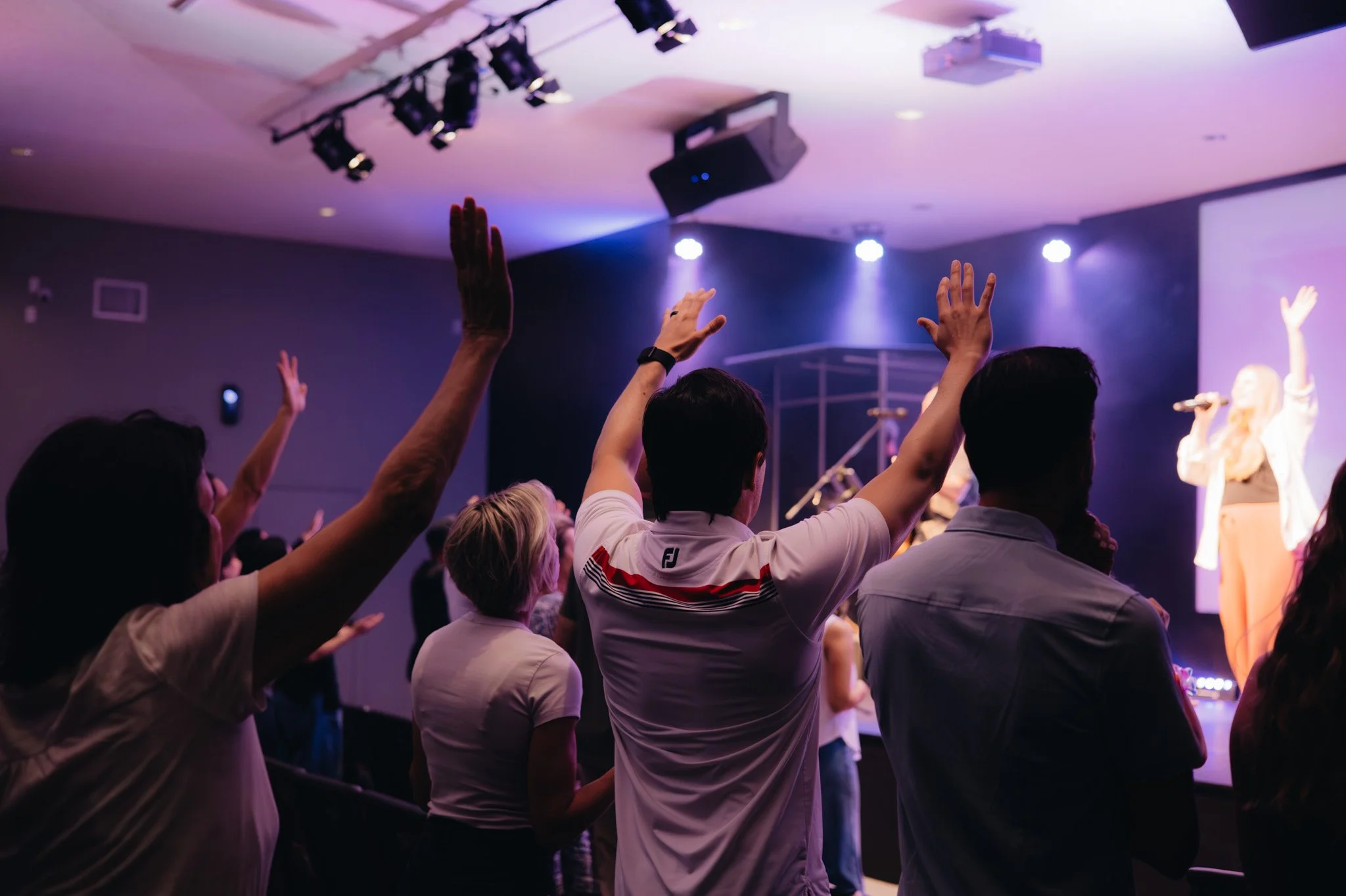 People raising their hands during a church gathering or worship event with stage lighting and a performer on stage.