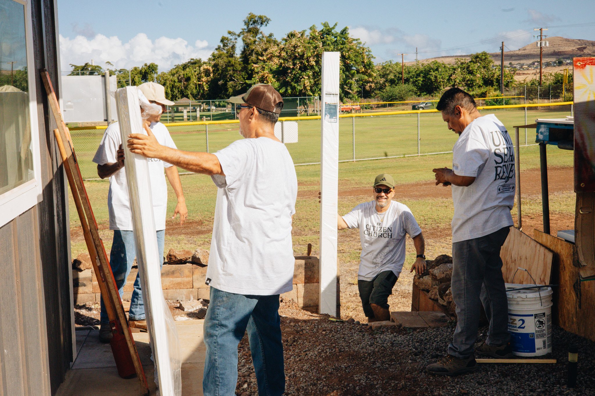 A group of people working together outdoors on a construction or landscaping project, with some holding a wooden or metal post and others standing nearby, on a grassy field under a partly cloudy sky.