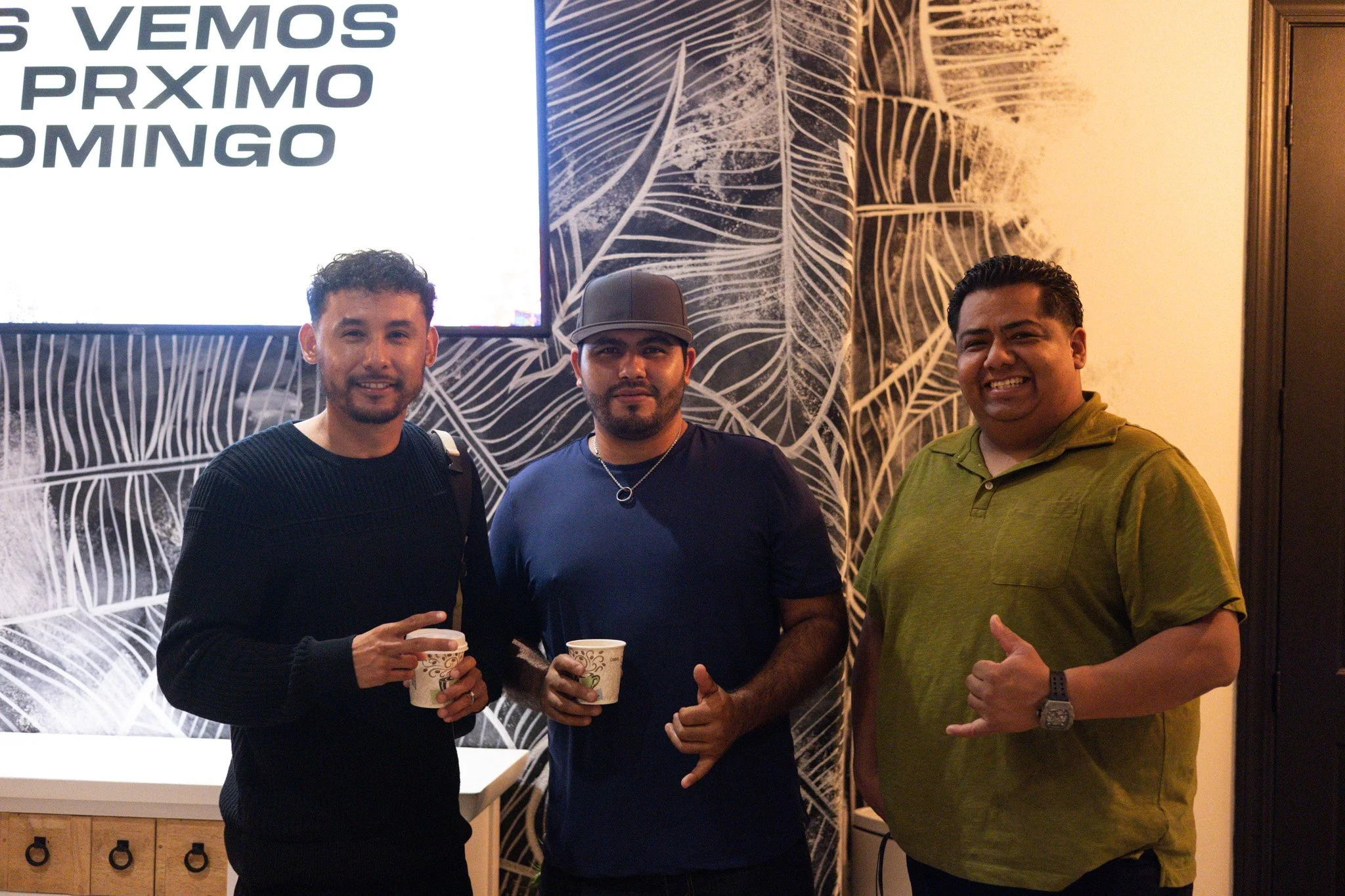 Two men sitting at an outdoor table, conversing and smiling, each with a drink in hand, wearing casual gray T-shirts and black caps, in a park-like setting with green grass and trees in the background.