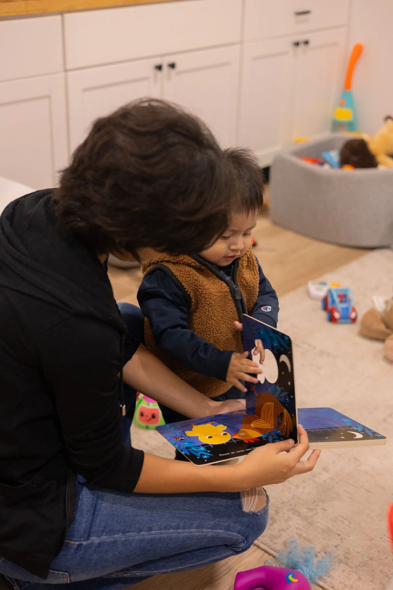 An adult helping a young child look at a children's pop-up book in a playroom.