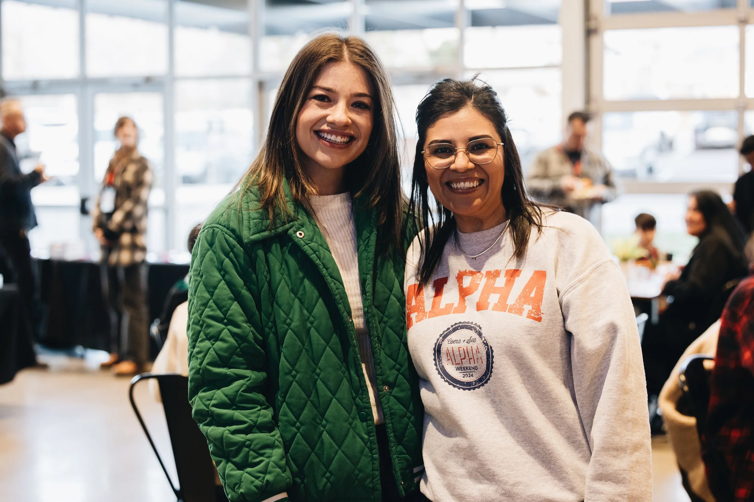 Two smiling women standing close together indoors, one wearing a green quilted jacket and the other in a white sweatshirt with orange lettering and a circular logo, at an event with other people in the background.