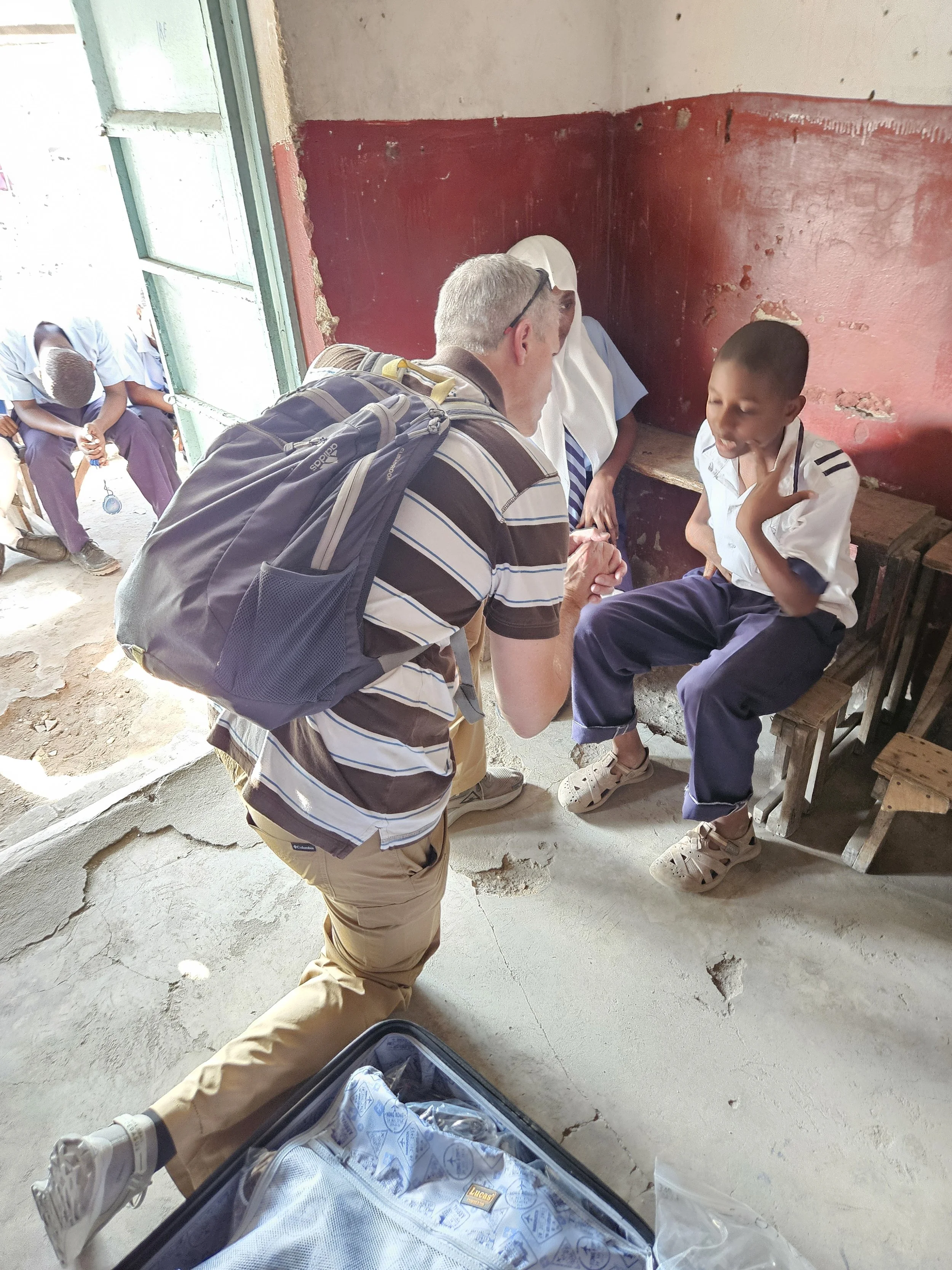 A man with a backpack kneeling and holding hands with a boy in a school uniform inside a classroom, with other children sitting outside near a door in the background.