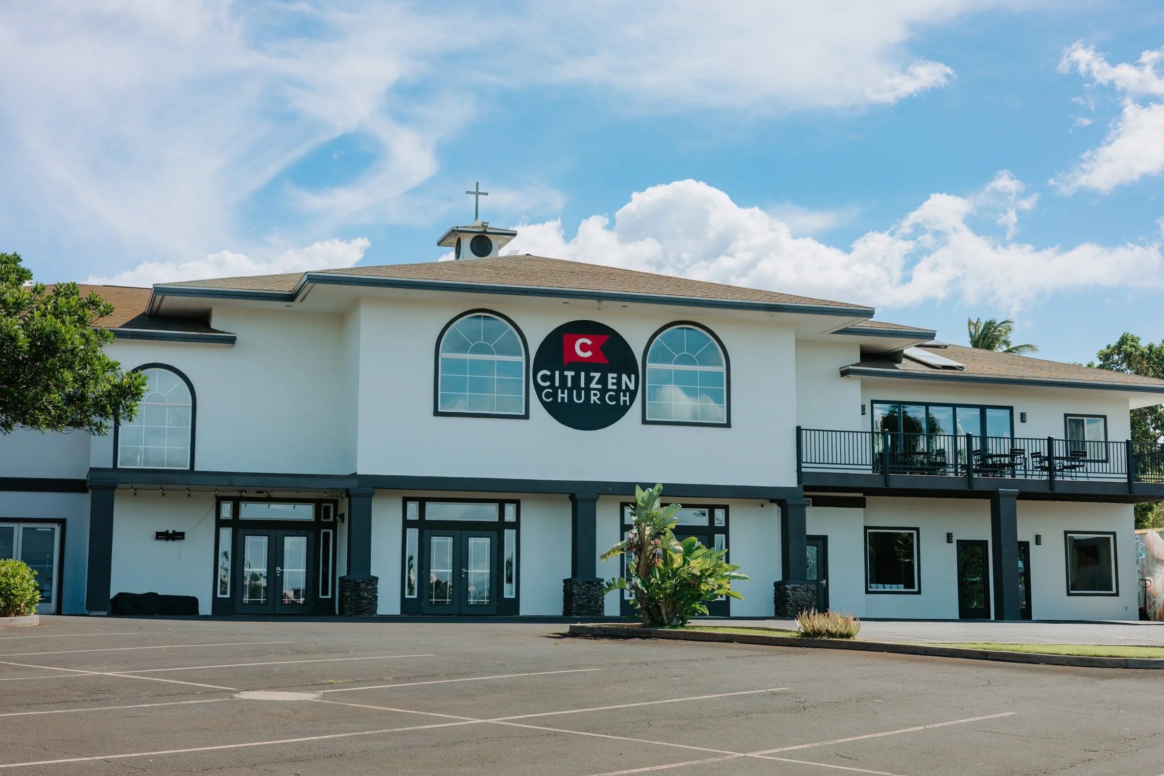 Exterior view of Citizen Church building with white walls, black framing, large arched windows, a parking lot in front, and a blue sky with clouds overhead.