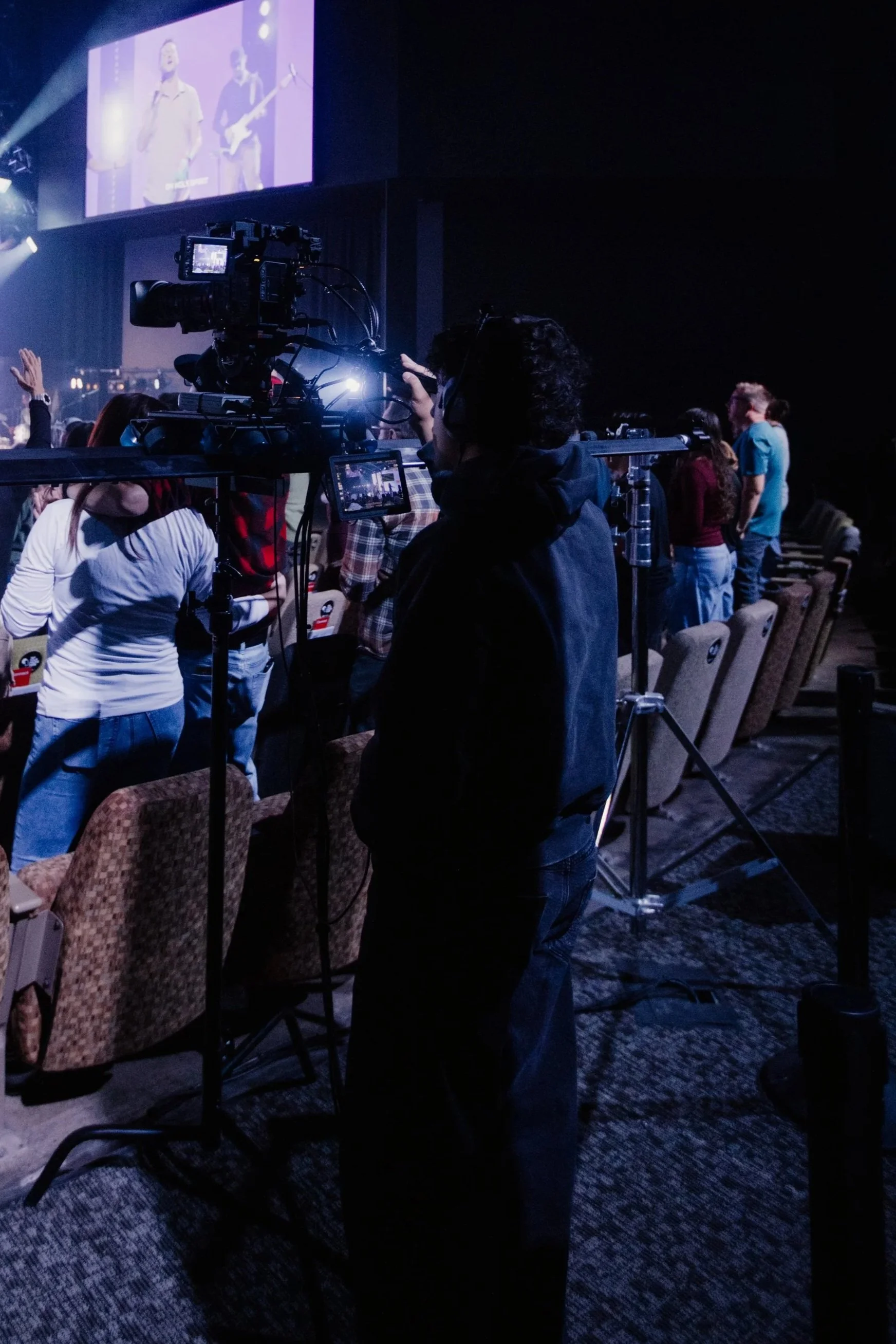 Photographer filming a concert on stage with large screen projection, audience standing and sitting in dimly lit auditorium.