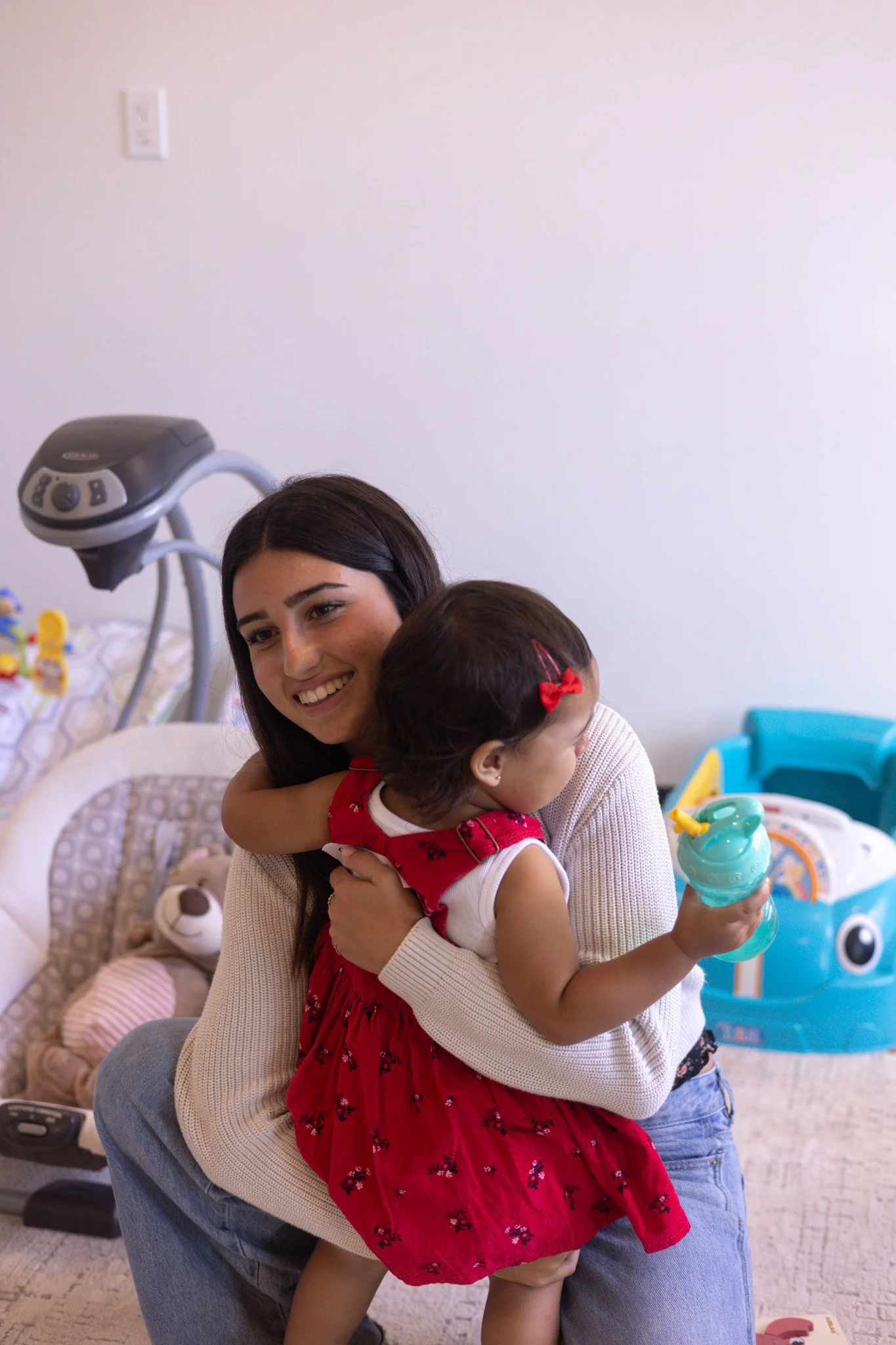 Woman holding a sleeping baby with a pacifier, sitting on a light gray chair in a room with blue walls.