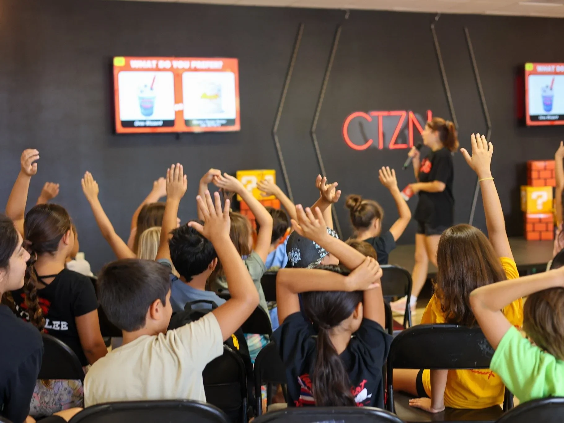 A group of children sitting in chairs, raising their hands while participating in an educational activity in a classroom or workshop setting. There is a woman holding a microphone at the front of the room, with two screens displaying questions or prompts in the background.
