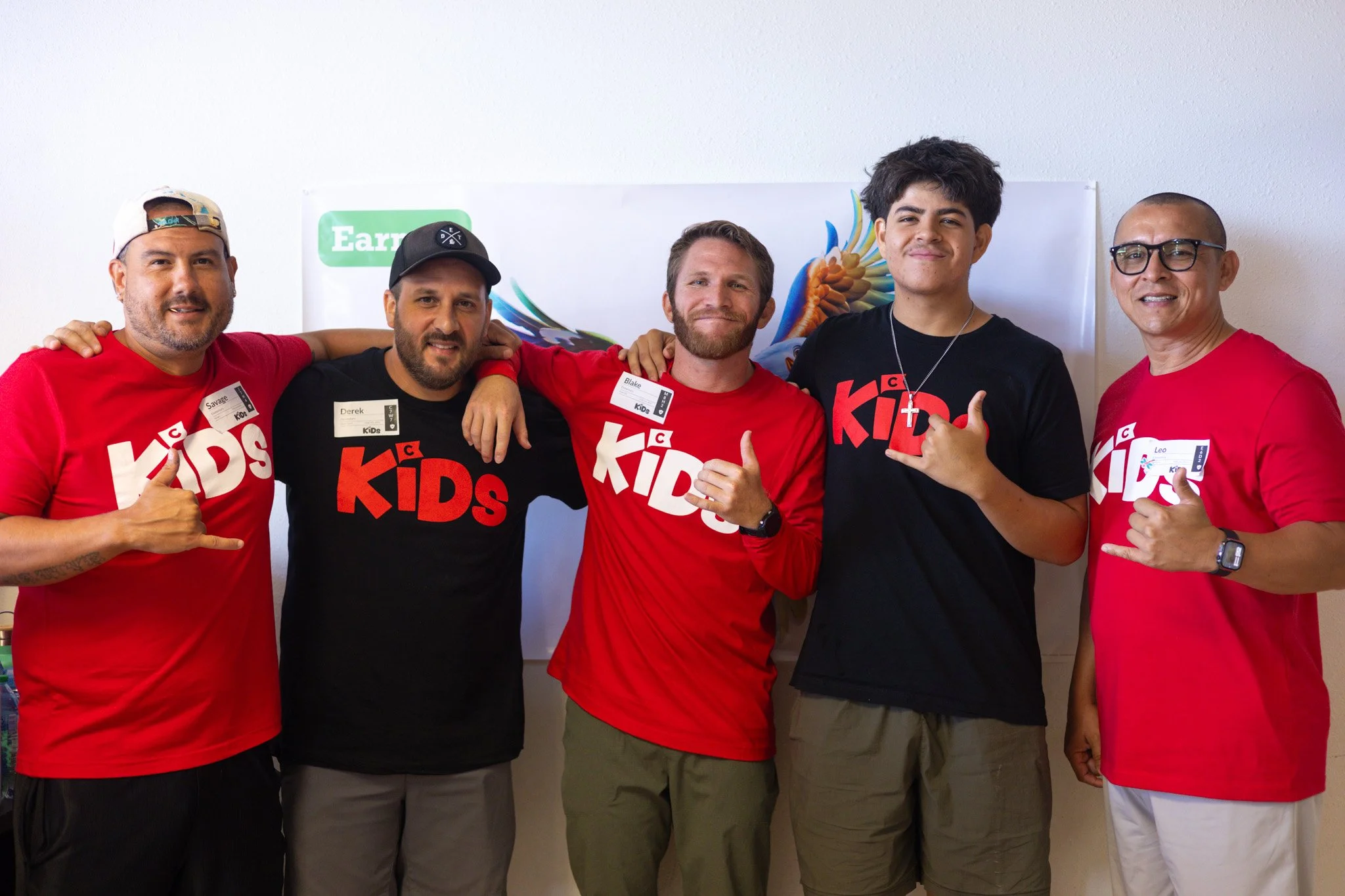 Five men standing together, all wearing red or black 'Kids' T-shirts, posing for a photo with their arms around each other. They are smiling and making hand gestures.