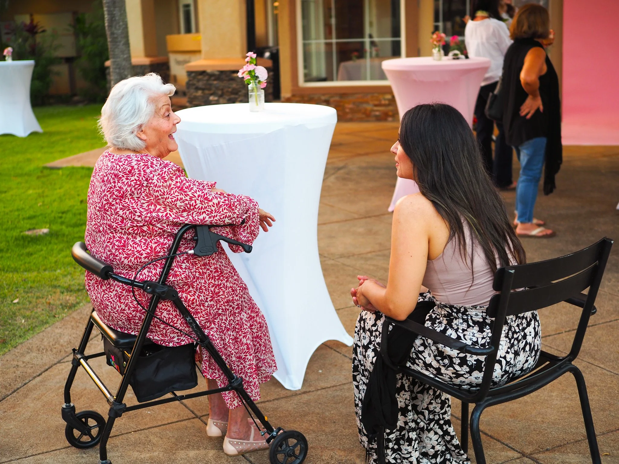 An elderly woman with white hair and a pink floral dress seated in a wheelchair is talking to a younger woman with long dark hair who is sitting on a black chair. They are outdoors at a social gathering with high cocktail tables and other people in the background.