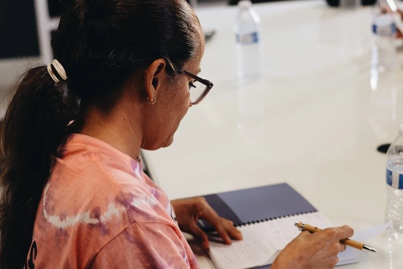 A woman with dark brown hair pulled back in a ponytail, wearing glasses and a pink tie-dye shirt, is sitting at a white table, writing in a notebook with a gold pen. There are water bottles on the table.