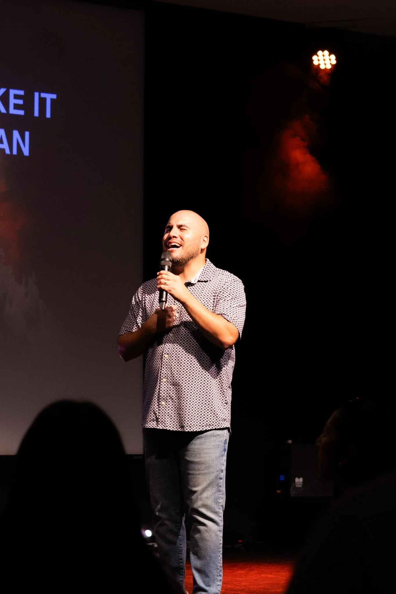 A man with a shaved head singing into a microphone on stage with a black background and red stage lights.