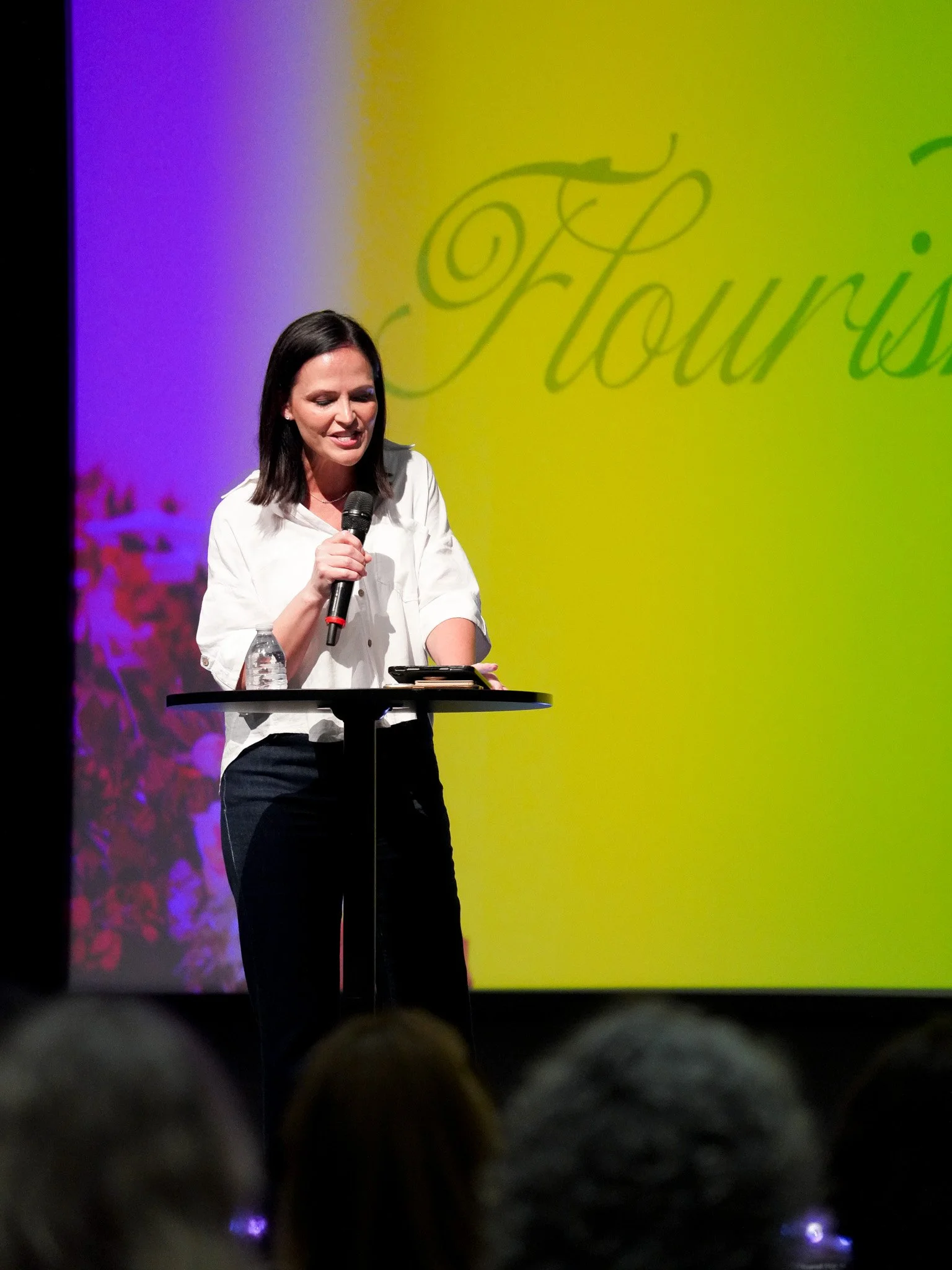 A woman with dark hair, wearing a white shirt, standing at a podium with a microphone. She is speaking or presenting in front of a colorful background with the word 'Touris' visible.