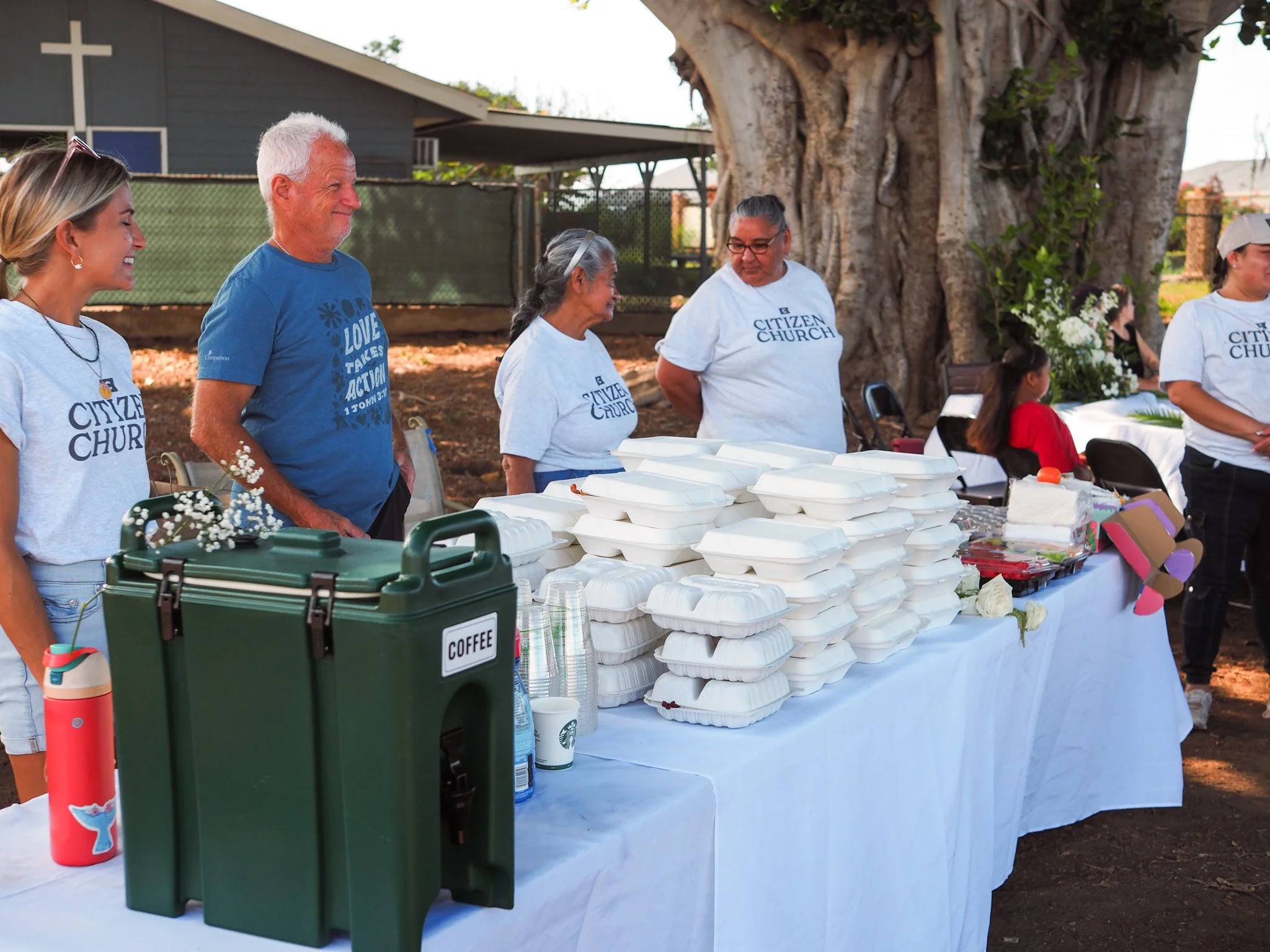 People standing behind a long table with stacked food containers, a coffee dispenser, and cups, at an outdoor event with a large tree in the background.