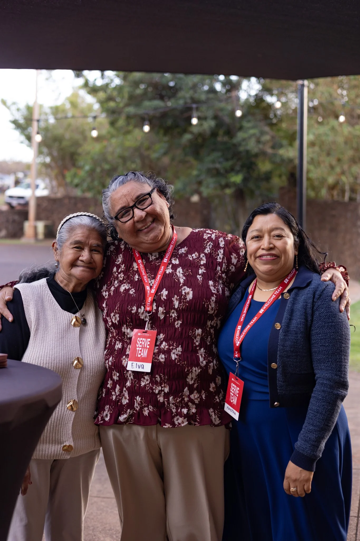 Three women smiling and posing outdoors at a social event, wearing name tags and lanyards, with string lights and trees in the background.