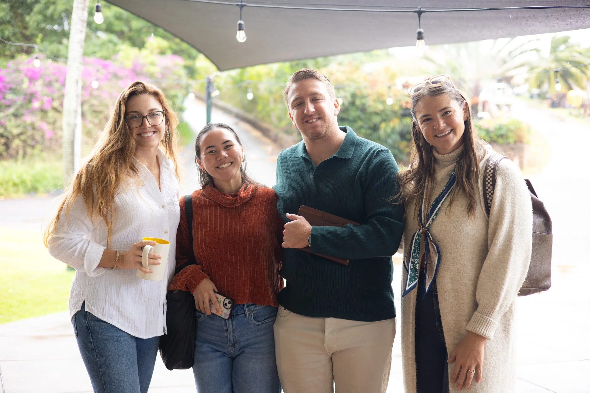 Four young adults standing together outdoors, smiling at the camera, with greenery and flowers in the background.
