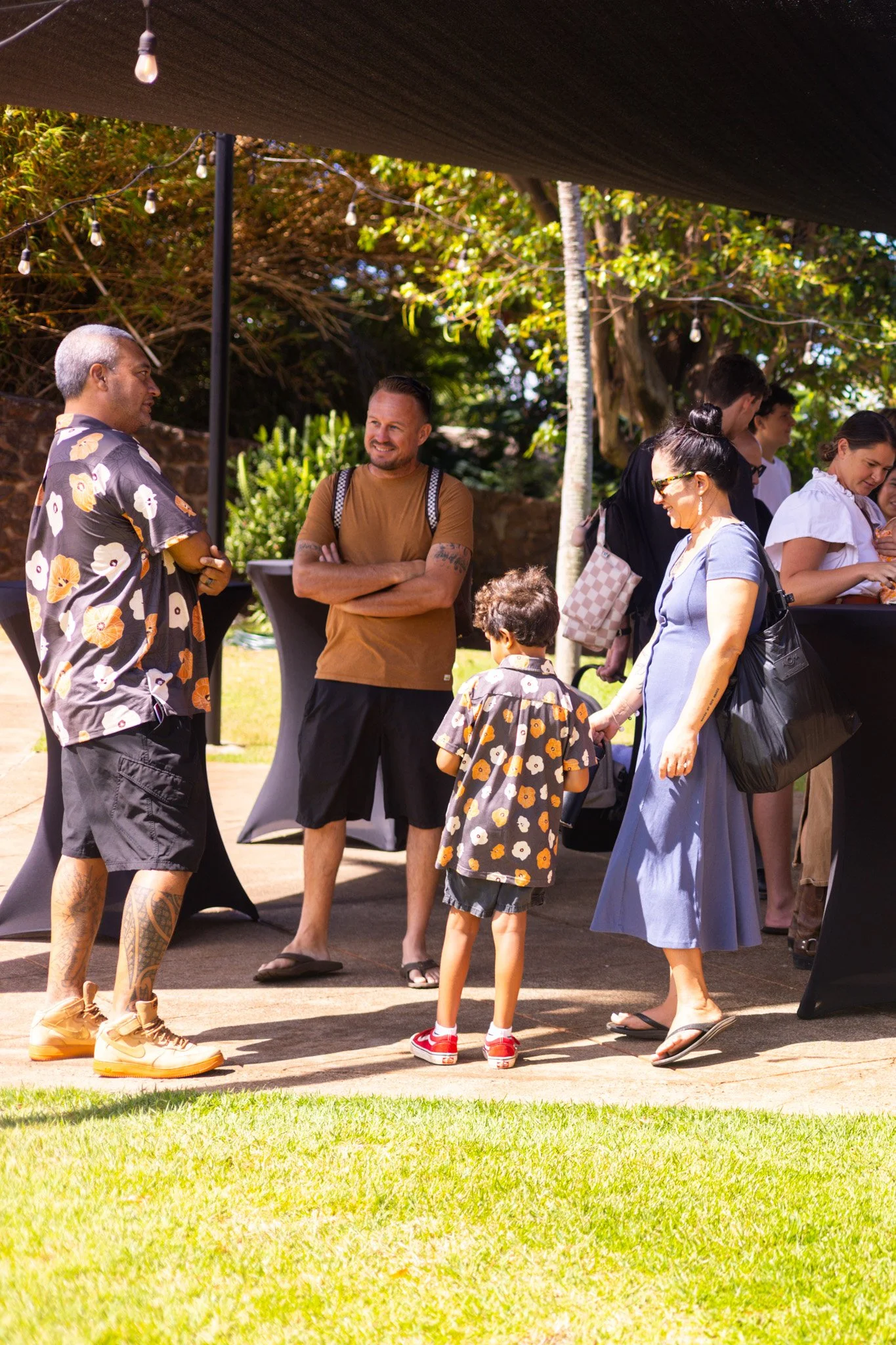 People socializing outdoors at a daytime event under a canopy, with a woman and child talking to a man wearing a floral shirt, and others standing nearby with tables and greenery in the background.