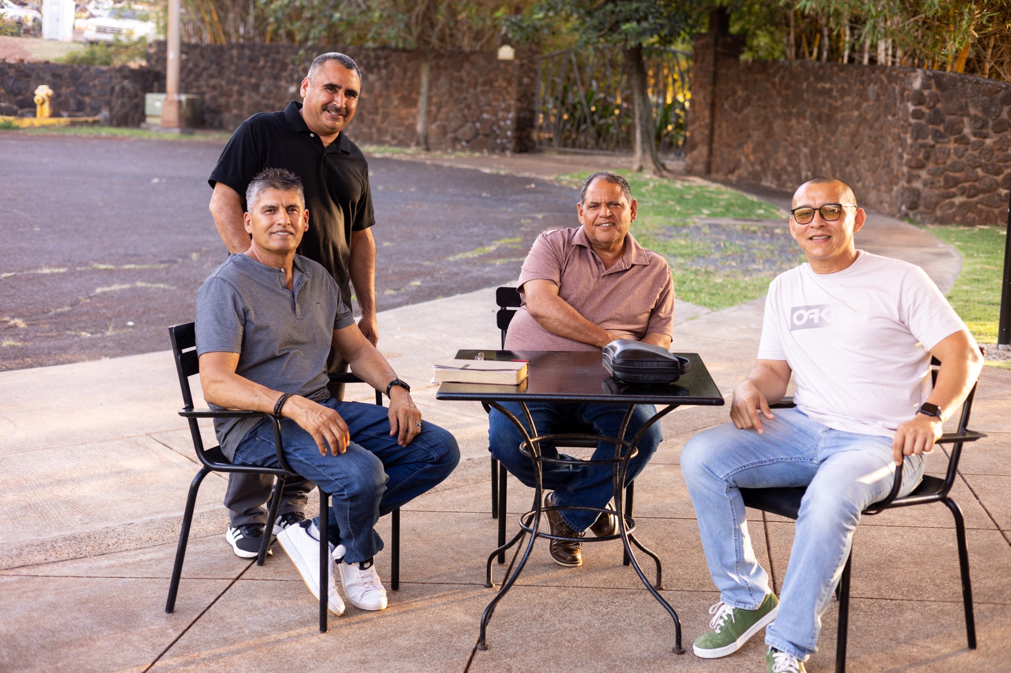 Four men gathered around an outdoor table, three sitting and one standing, with books and a black bag on the table, in a park or plaza.