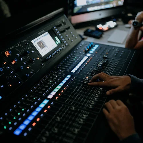 Close-up of a professional audio mixing console with illuminated controls, a person operating it, and a monitor in the background.
