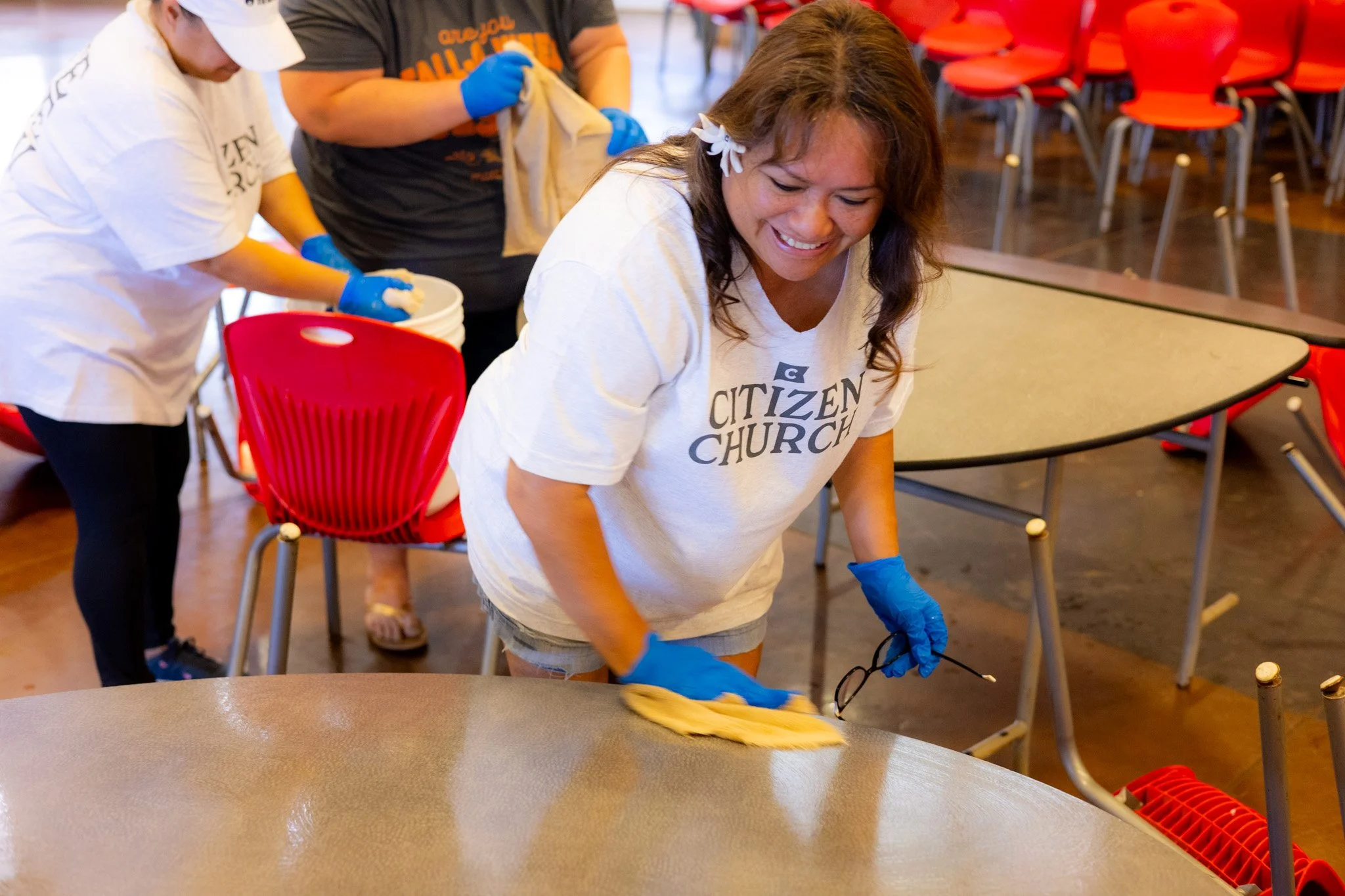 Women cleaning tables inside a room for community service, wearing gloves and casual t-shirts, some with 'Citizen Church' logo.