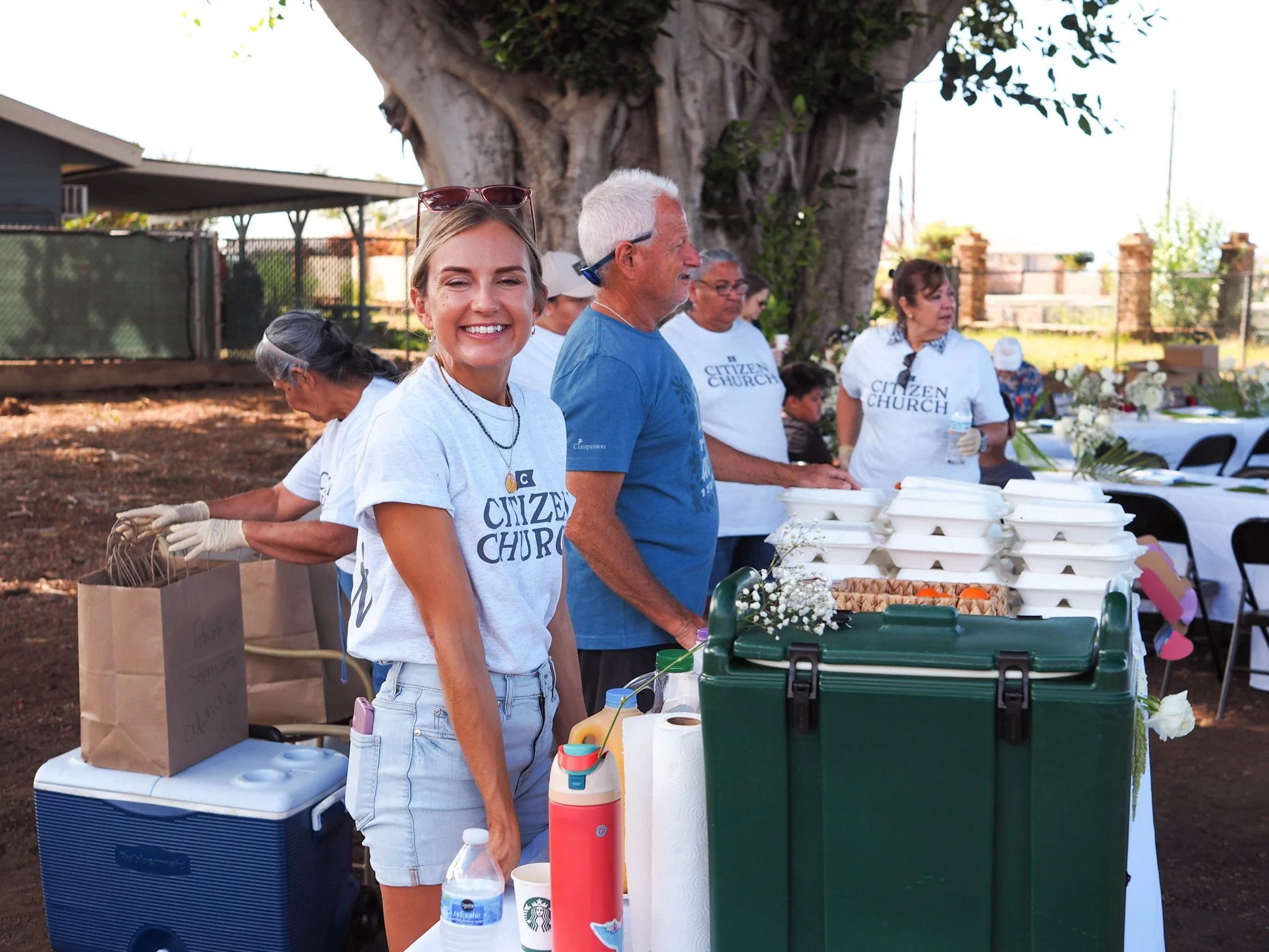 Group of people at a community event serving food outdoors, with tables, containers, and a large tree in the background.