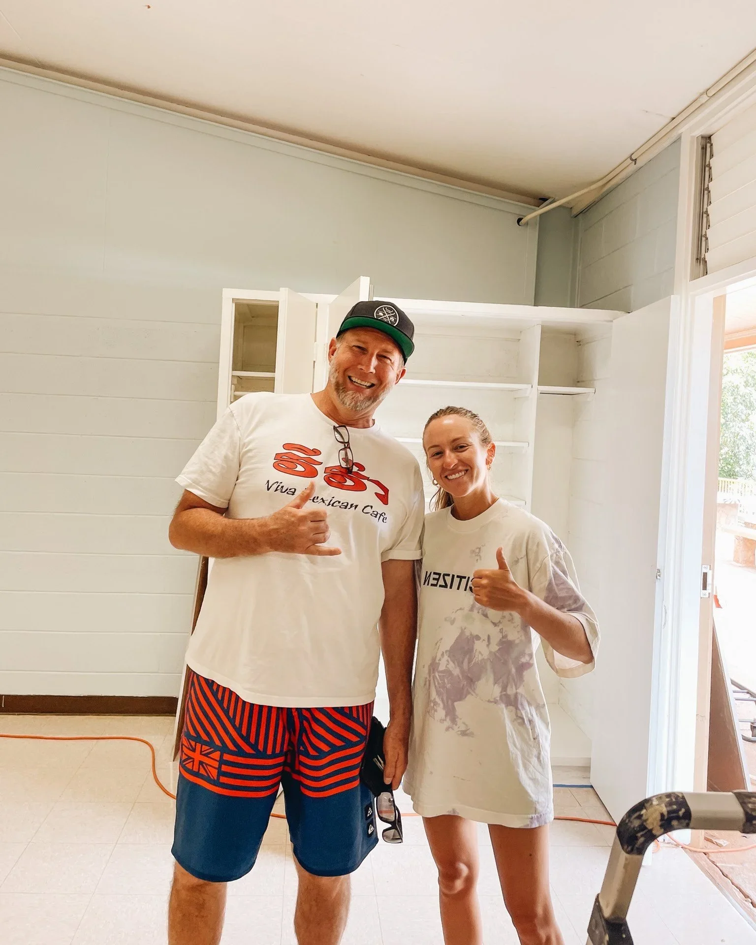 Two smiling people, a man and a woman, standing indoors in front of a white wall with open cabinets. The man is wearing a white t-shirt, colorful striped shorts, and a baseball cap. The woman is wearing an oversized T-shirt and giving a thumbs-up. They appear to be happy, possibly in a home renovation or construction setting.