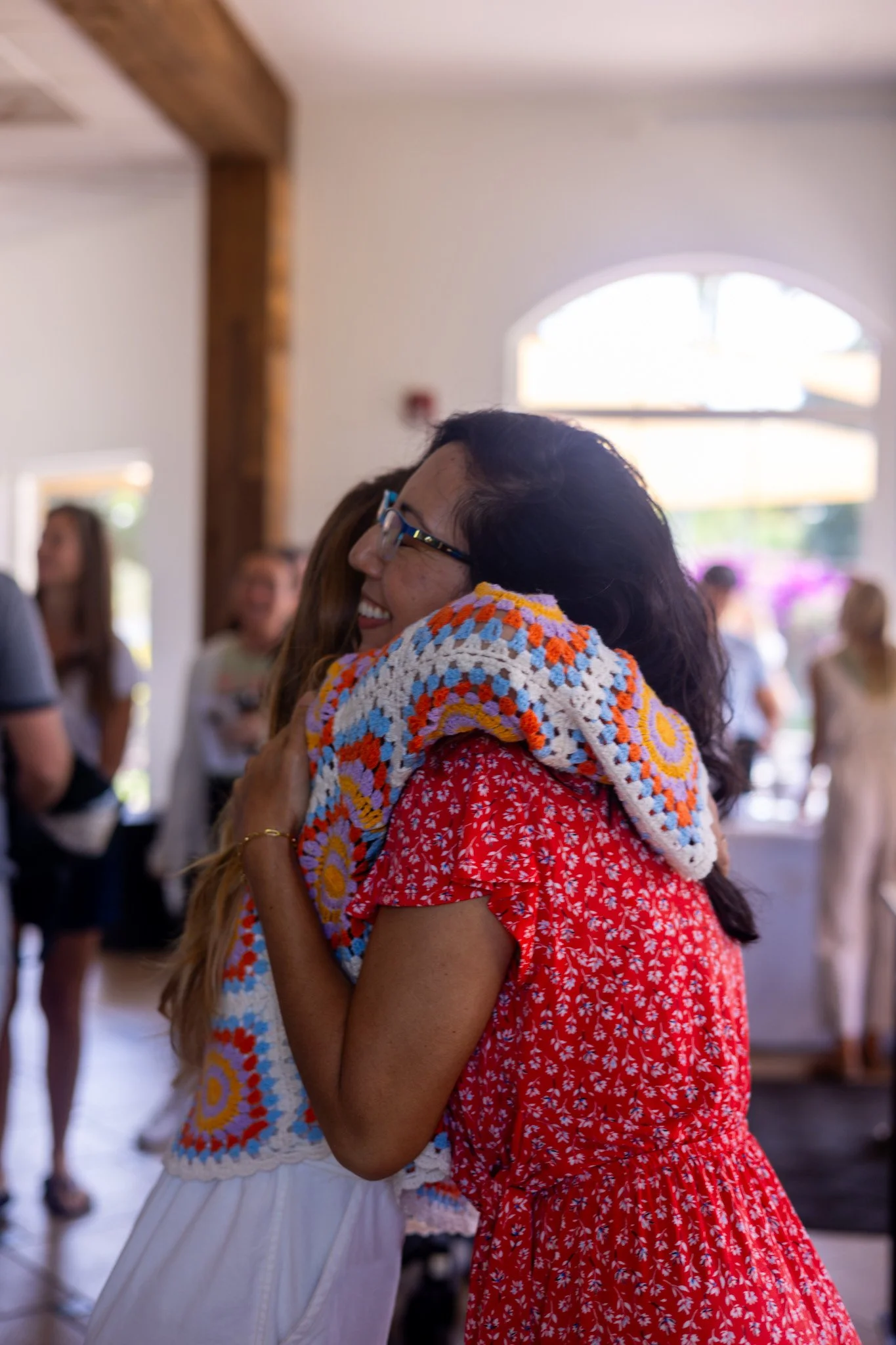 Two men sitting at an outdoor table, conversing and smiling, each with a drink in hand, wearing casual gray T-shirts and black caps, in a park-like setting with green grass and trees in the background.