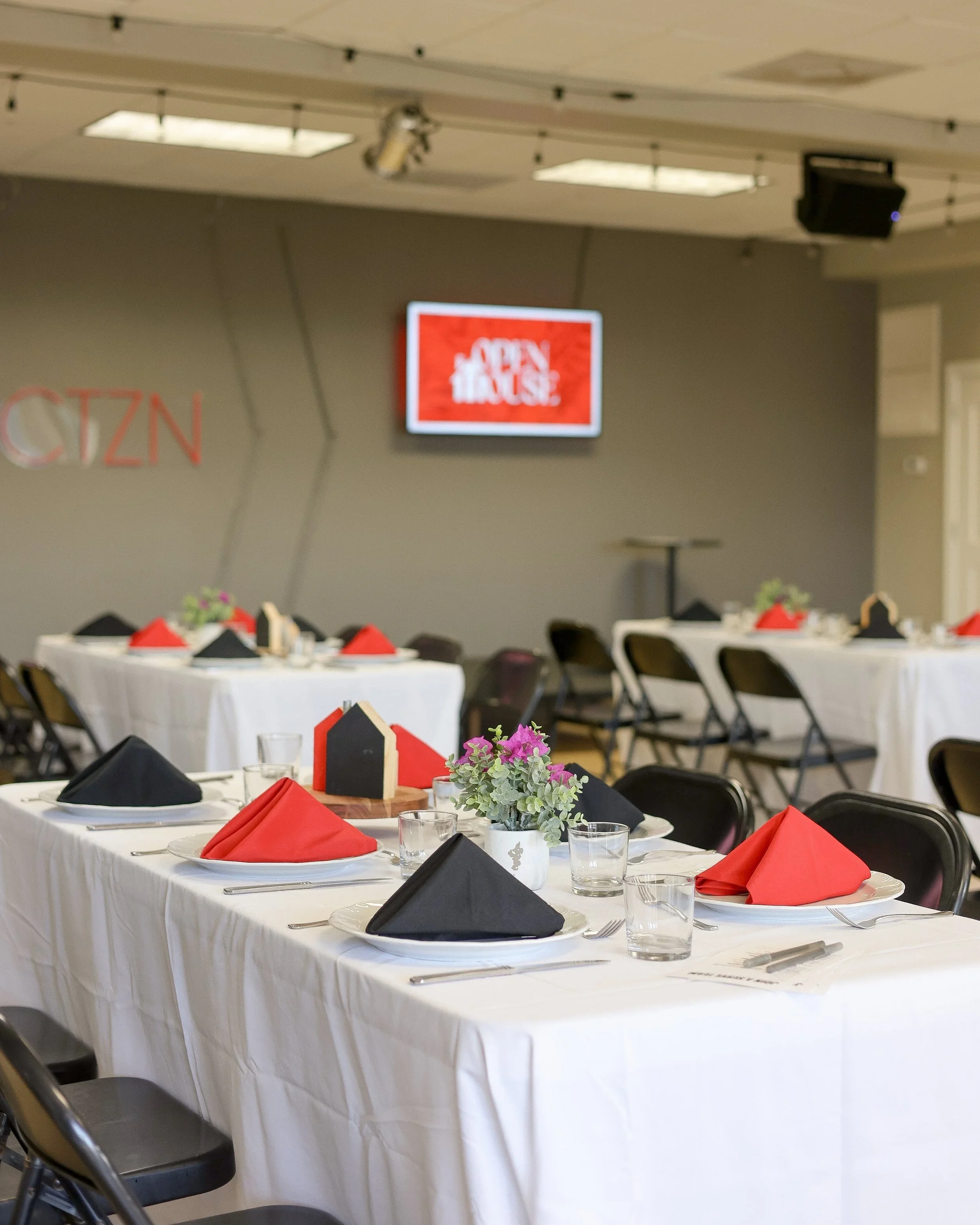 Dining hall with round and rectangular tables set with white tablecloths, black and red napkins, glasses, cutlery, and small potted plants with purple flowers, in front of a gray wall with a red and white sign that says 'Open House' and a television screen.
