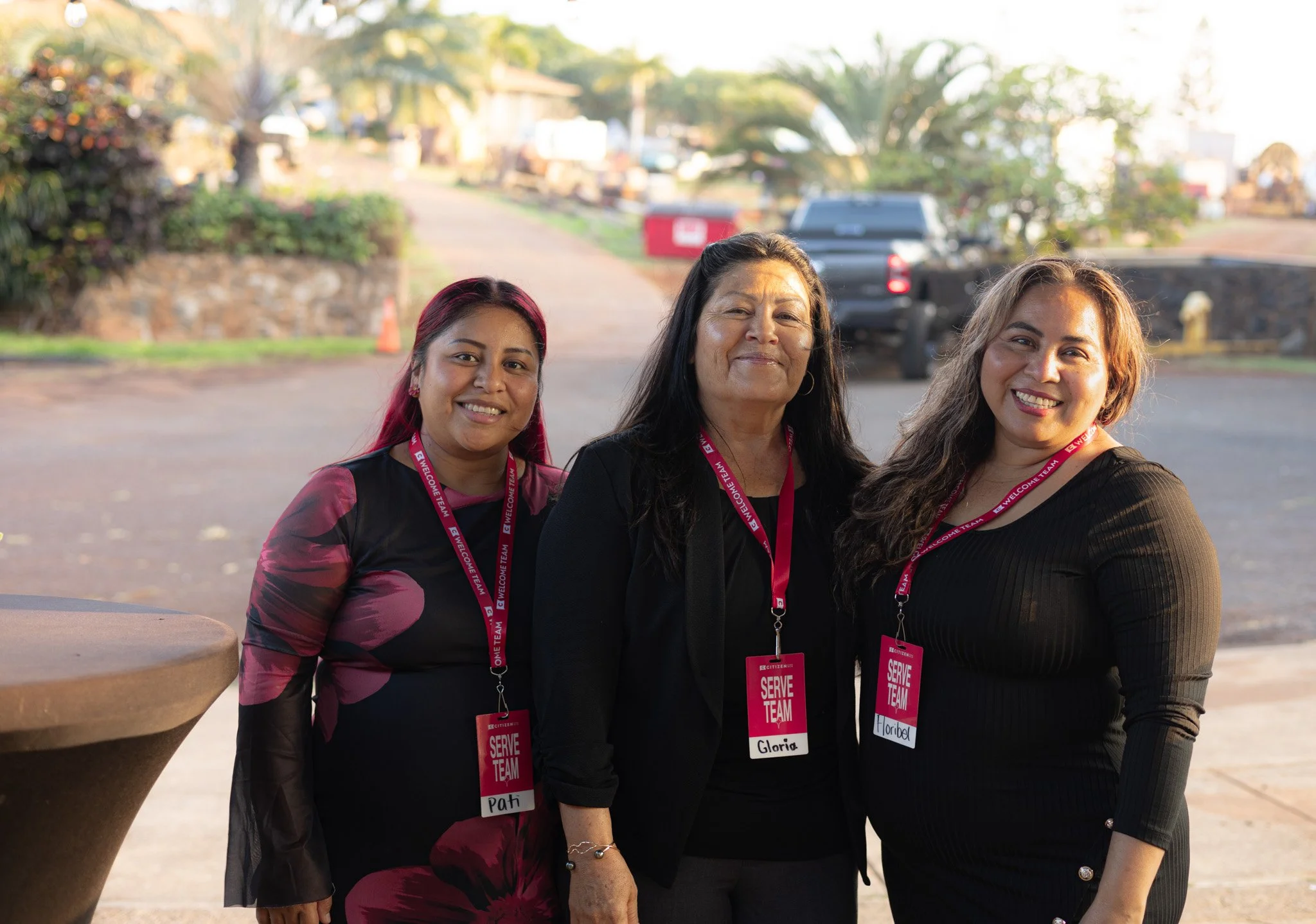 Three smiling women with event lanyards and badges standing outdoors on a street or parking lot, with trees and vehicles in the background.