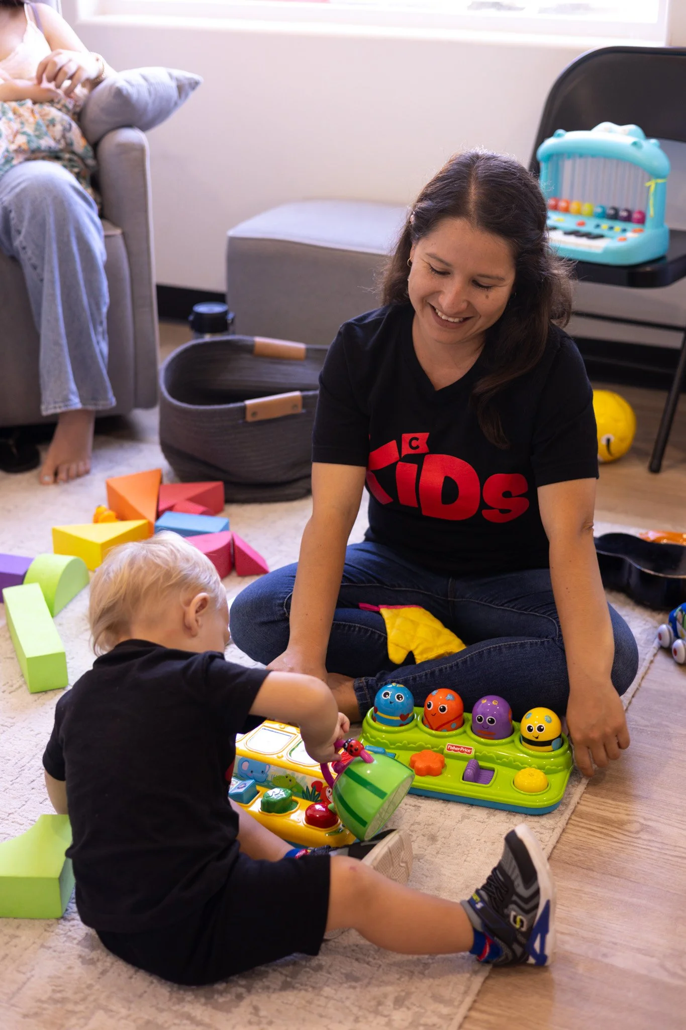 A woman and a young boy playing with colorful children's toys on the carpeted floor of a living room, with various toys and furniture visible in the background.
