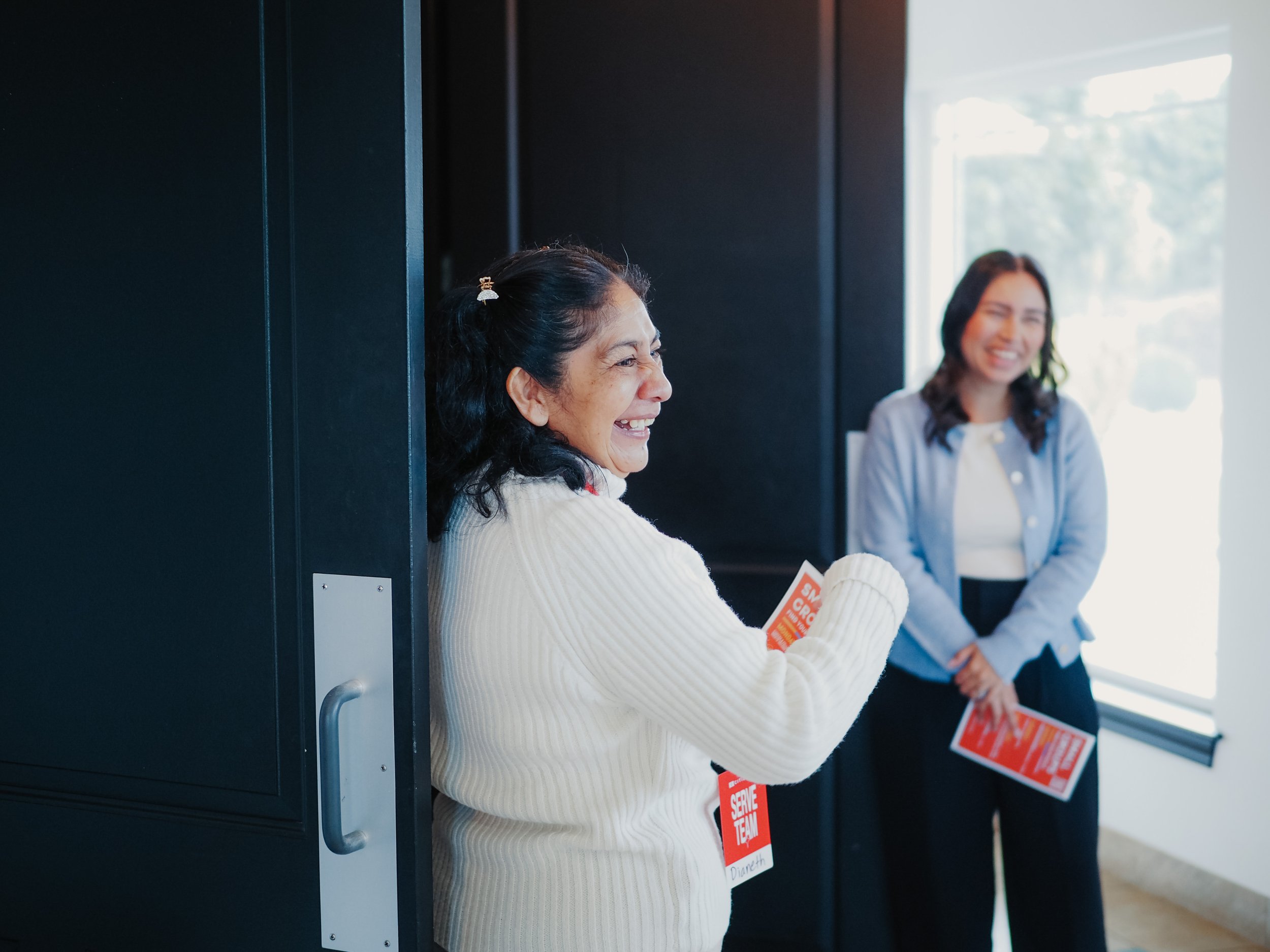 Two women laughing and holding red signs or pamphlets, one standing in a doorway and the other near a window, in a bright indoor setting.