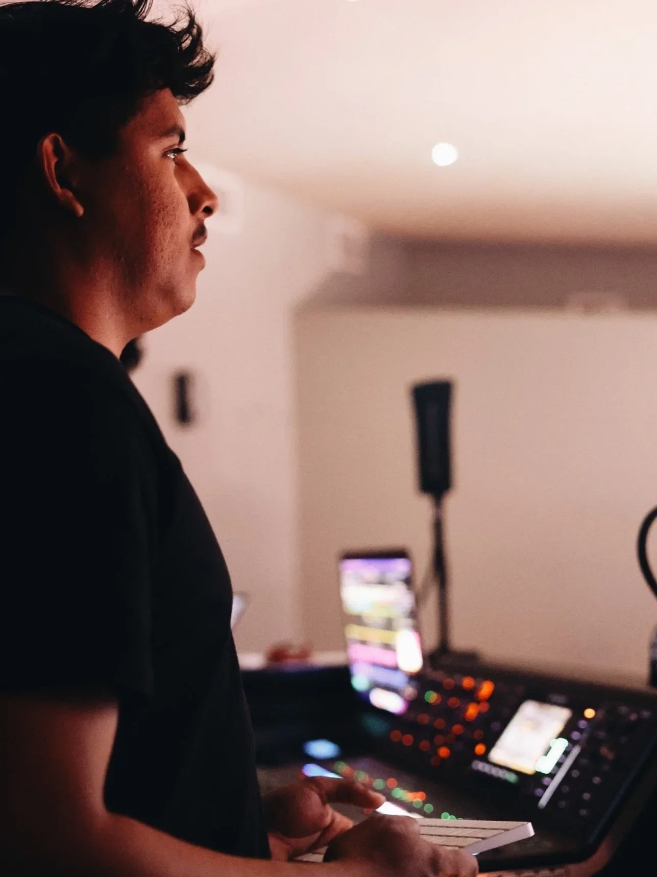 A young man in profile standing in front of DJ equipment, holding a notebook, with a monitor and speakers in the background.