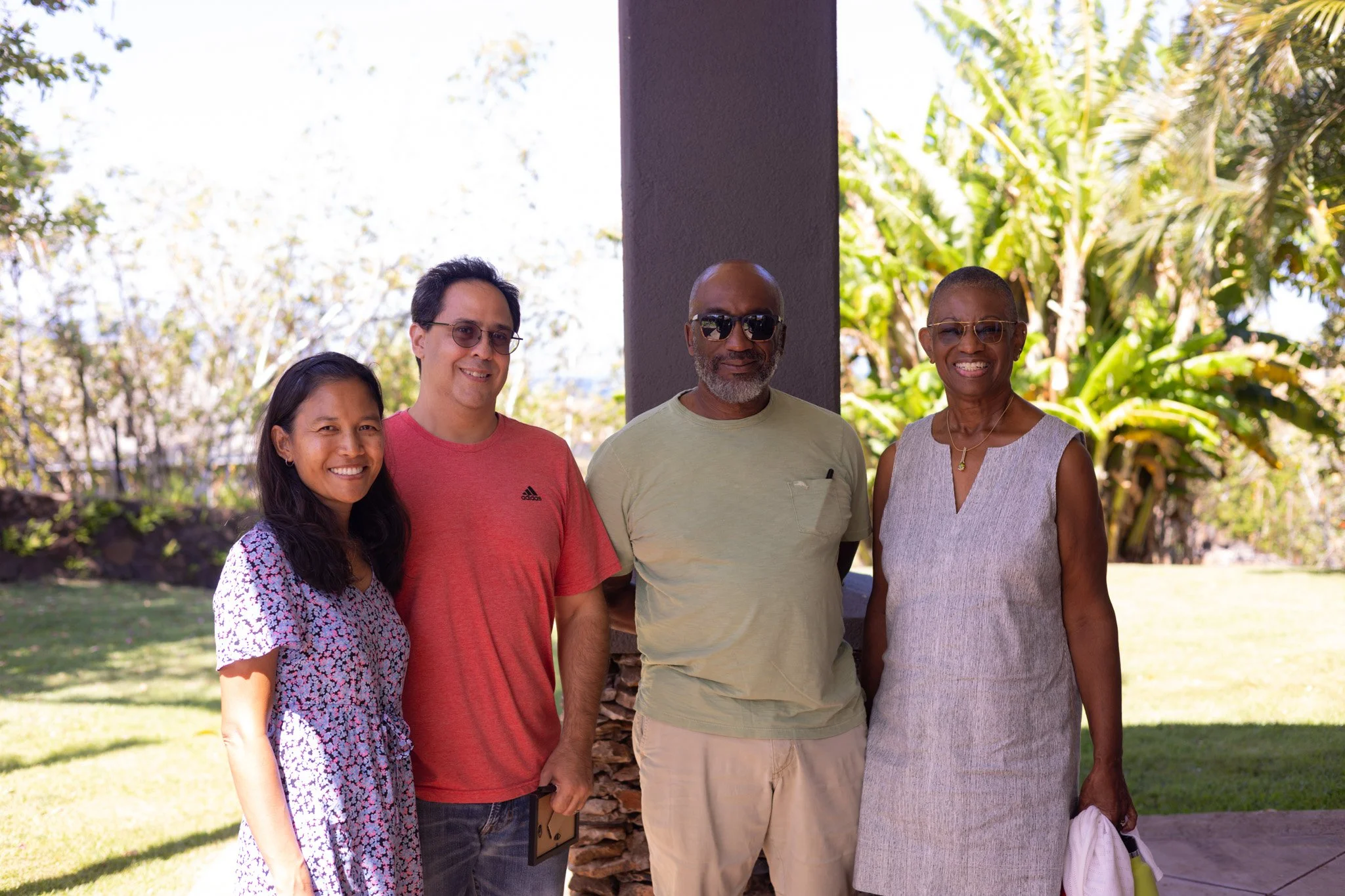 Four people standing outdoors in front of a tree and greenery on a sunny day.