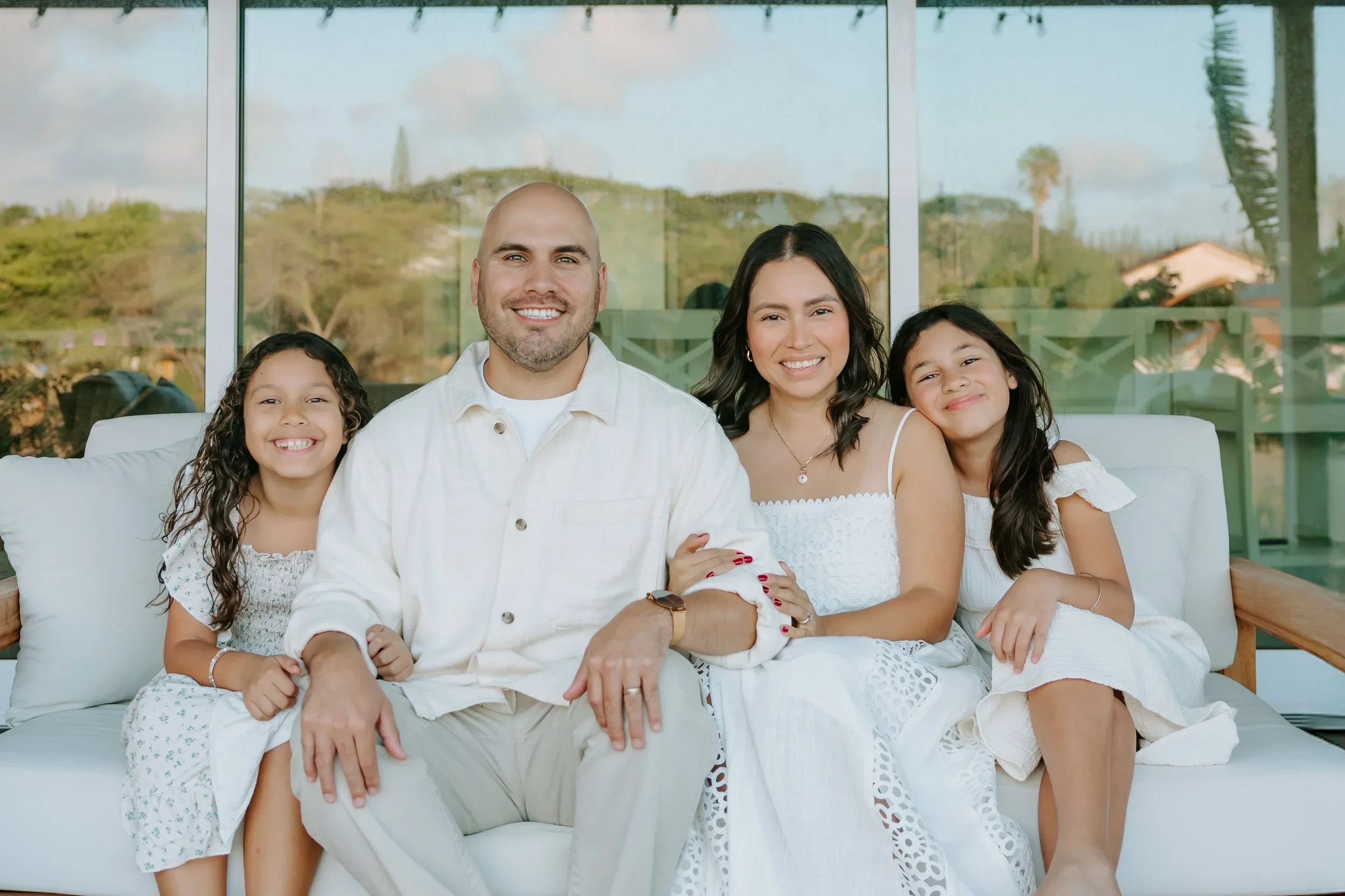 Family of four sitting on a white couch indoors, with large windows showing a green, hilly landscape outside. The father is bald with a beard, wearing a white shirt. The mother has dark hair, wearing a white dress. Two young girls with long, dark hair are sitting beside them, all smiling.