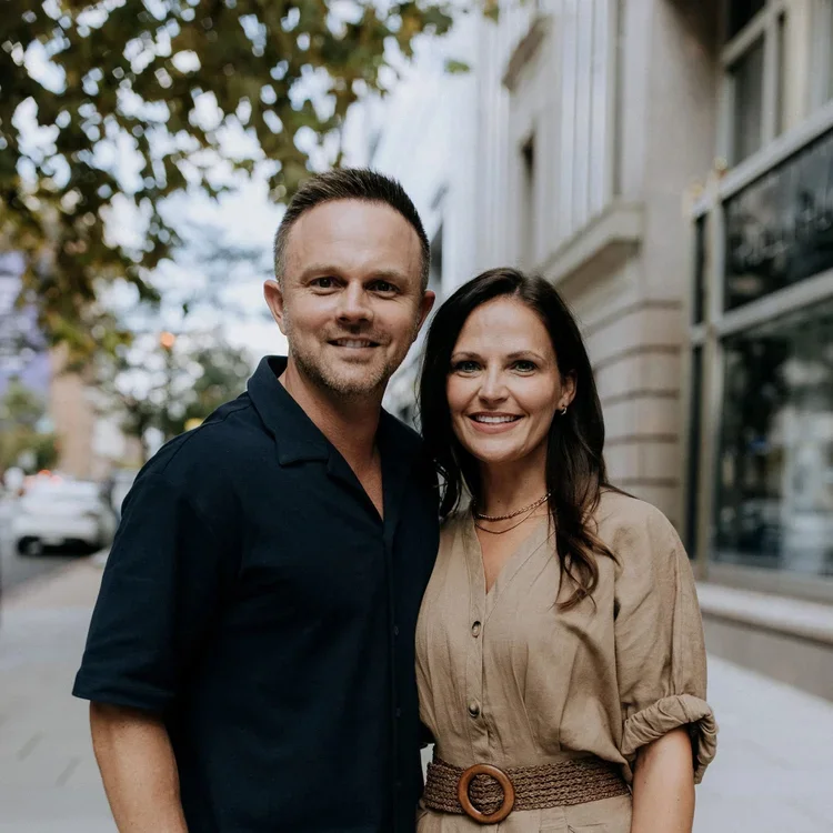 A smiling man and woman standing outdoors on a city sidewalk, with trees and building storefronts in the background.