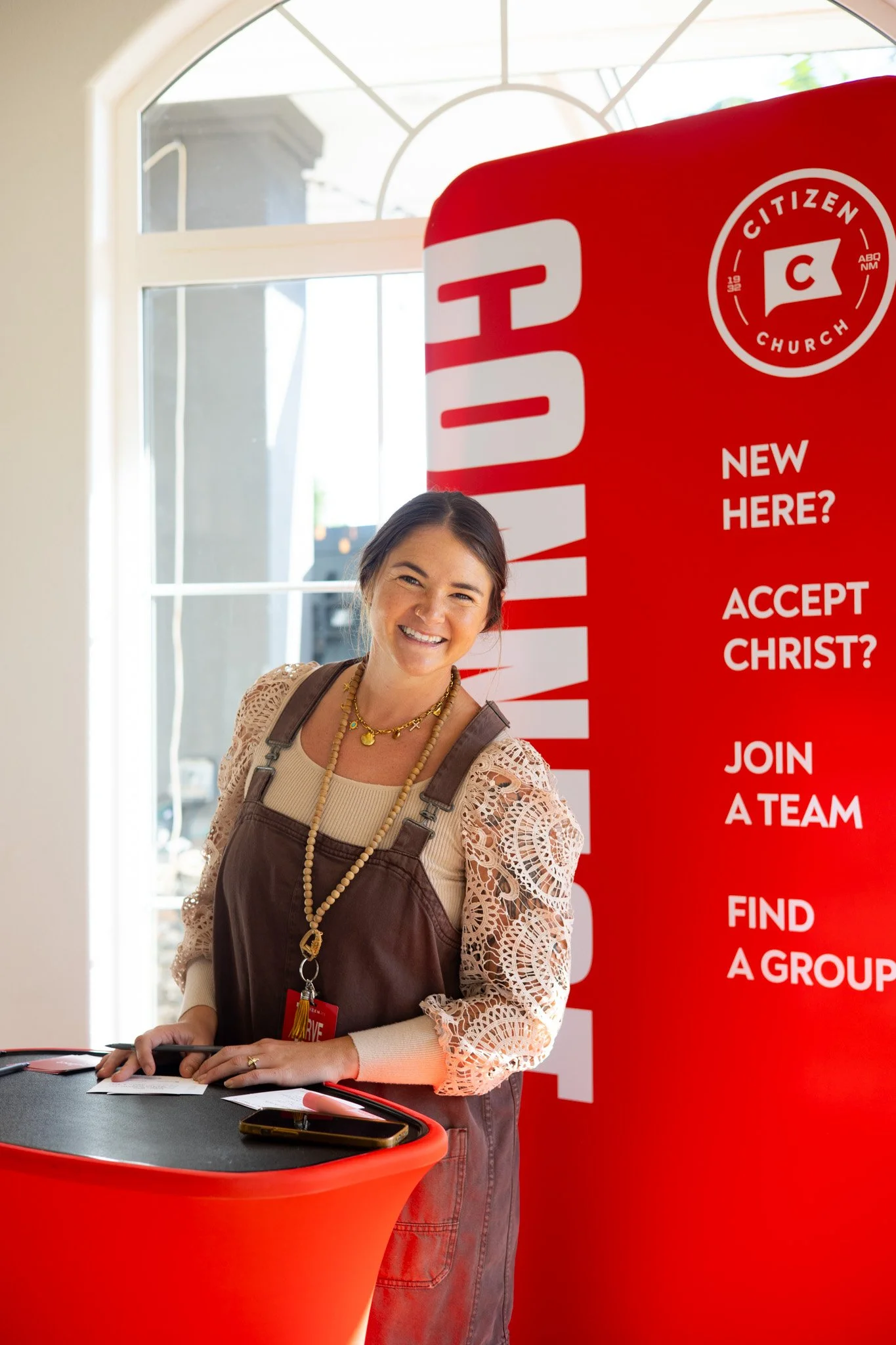 A smiling woman standing at a red registration desk with a background of large windows, a red banner with white text about joining a church, and sunlight coming through the windows.