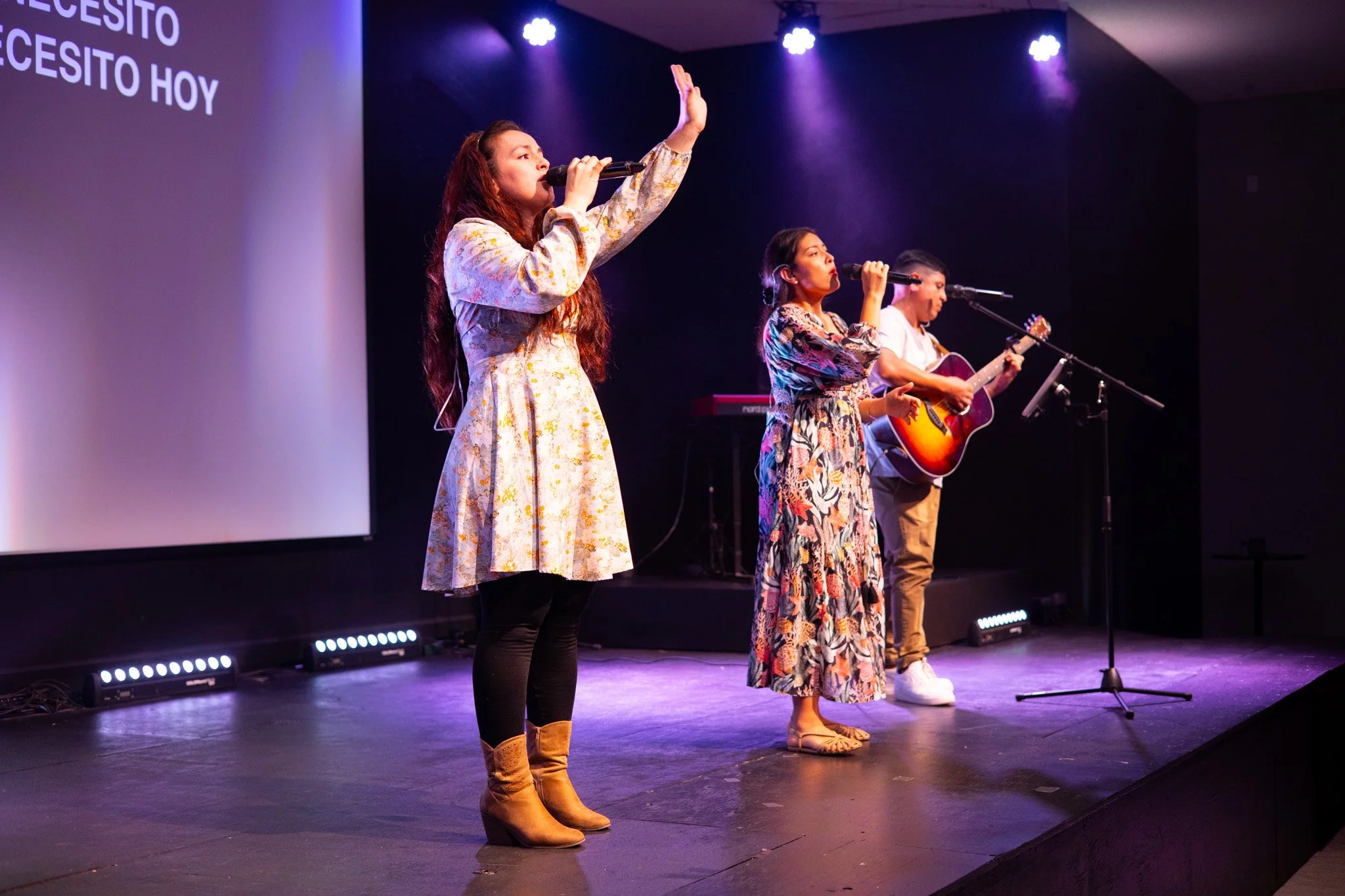 Three women performing on stage with microphones, one playing guitar, in a sanctuary-like setting with stage lighting.