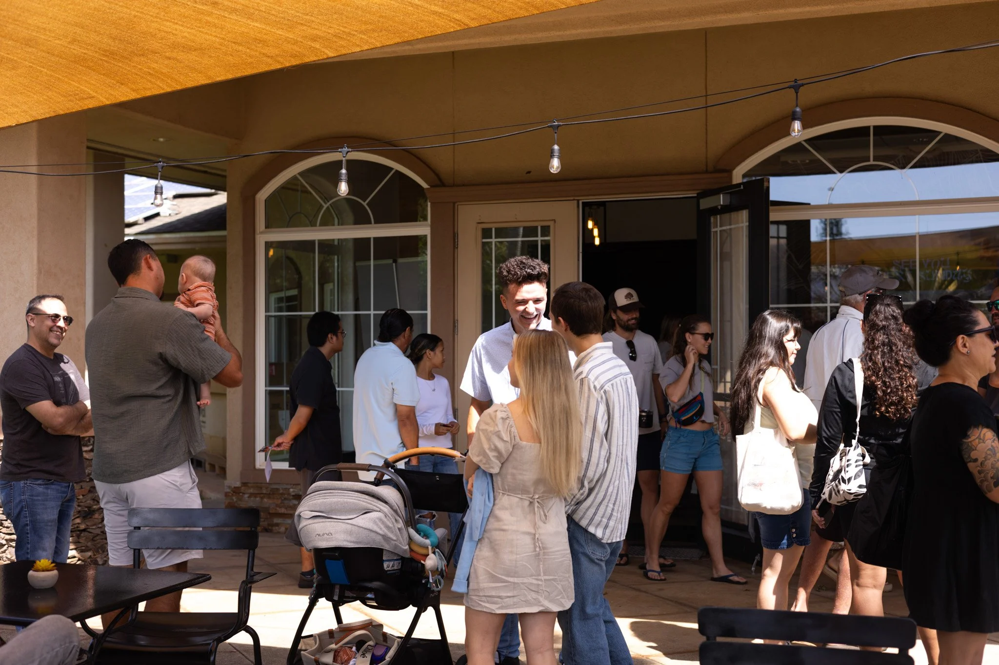 Group of people socializing outside a house, some engaged in conversation, some waiting in line, with a stroller in the foreground.