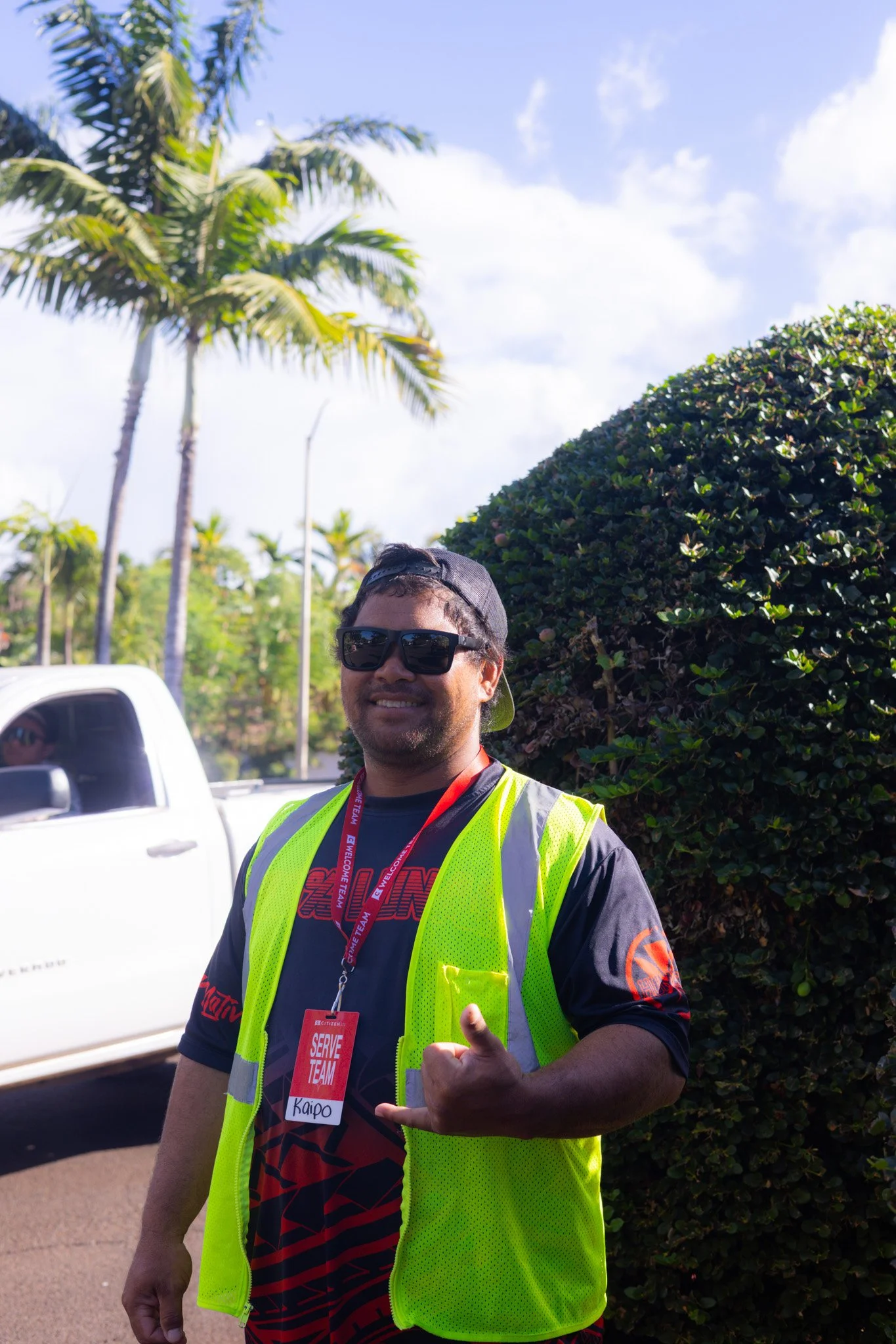 A man wearing sunglasses, a black cap backwards, a bright yellow safety vest, and a black shirt with red and orange design, smiling and pointing with a shaka sign, standing outdoors near a white truck and green foliage under a partly cloudy sky.
