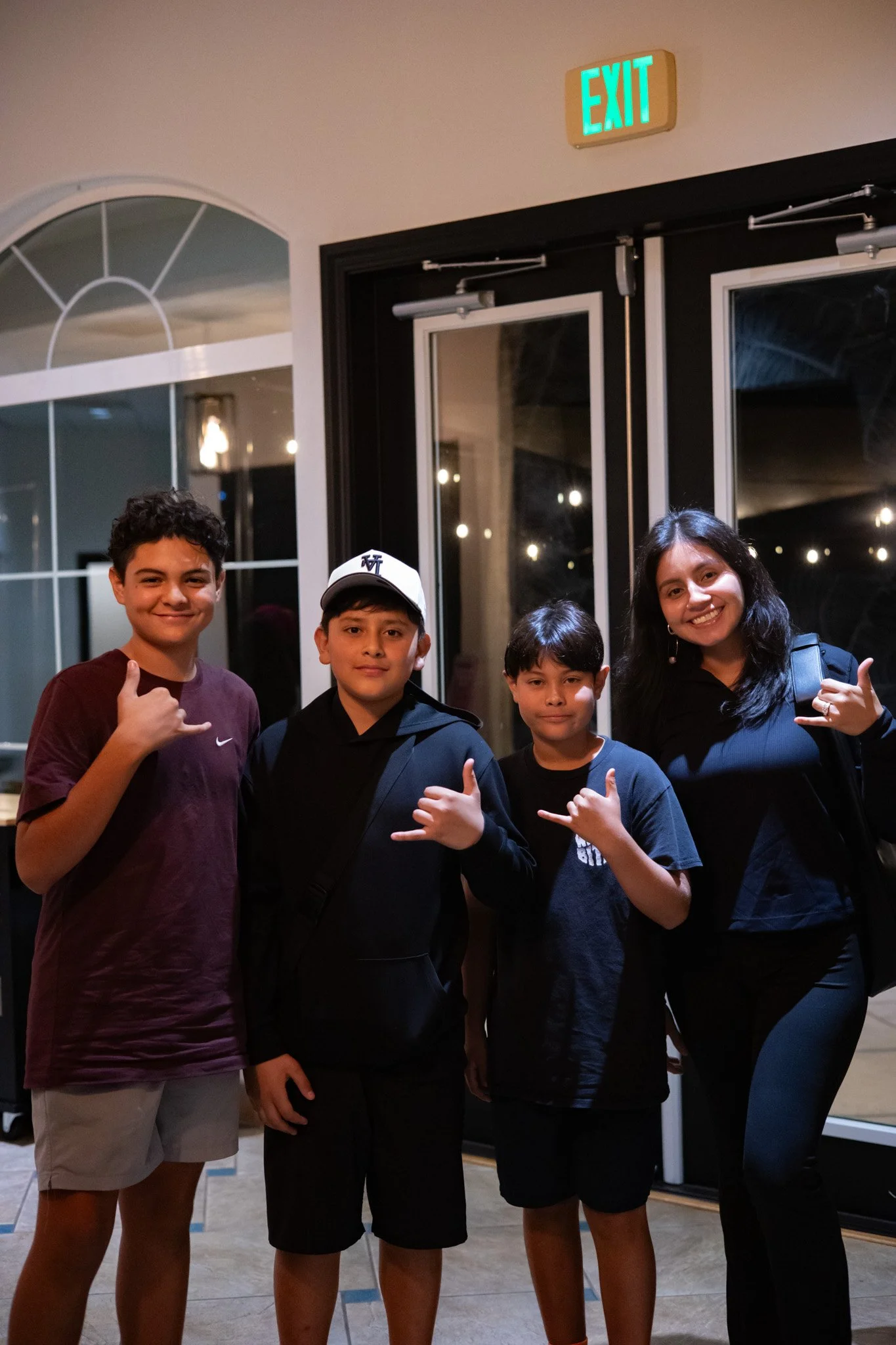 A group of four children and one adult woman standing together inside a building near a door with an illuminated exit sign, posing for a photo and making shaka hand gestures.