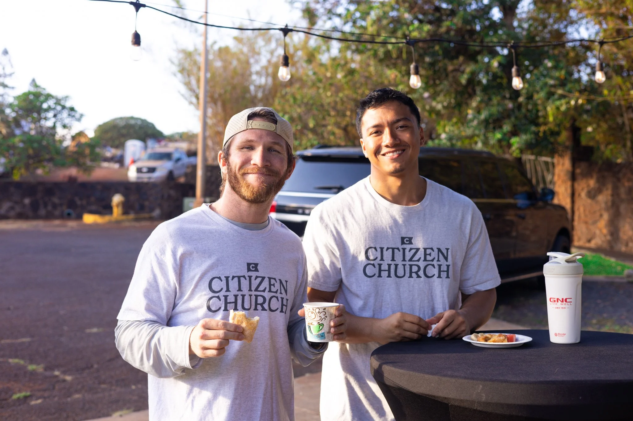 Two young men stand outdoors at sunset, smiling, wearing gray Citizen Church T-shirts, holding food and drinks, with string lights and parked cars in the background.