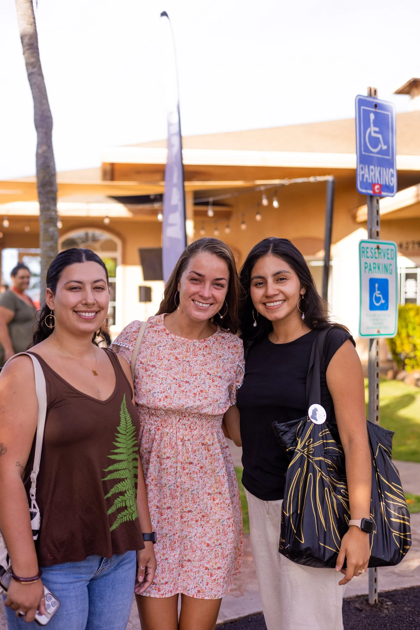 Three women smiling and posing together outdoors, near parking signs for handicapped accessible parking.