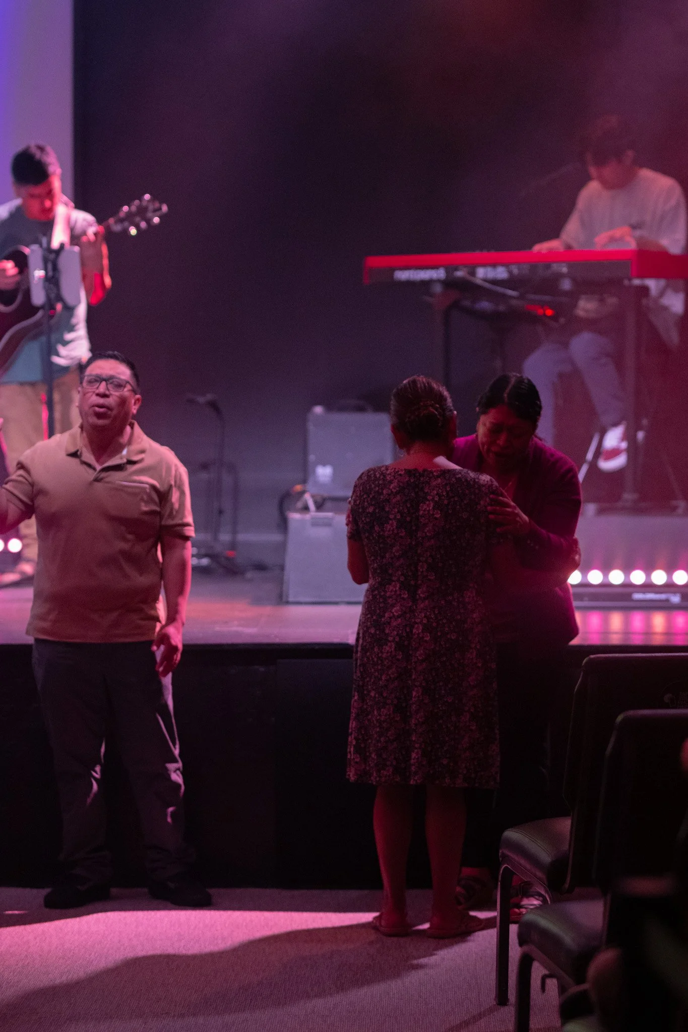 People on stage during a performance or event, with two individuals in front of the stage, one singing or speaking and another holding their shoulders, and two musicians in the background playing instruments on a dark, lit stage.