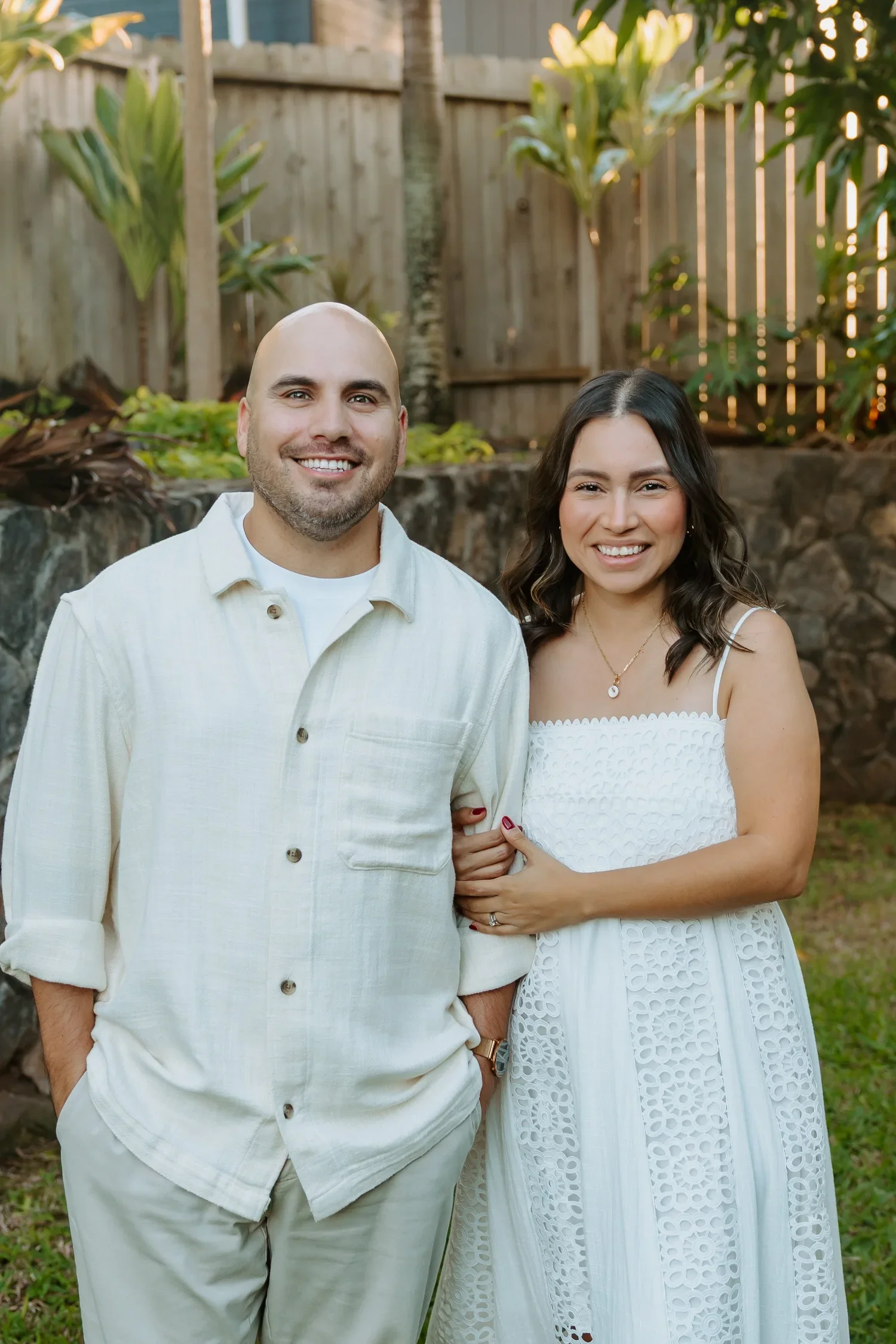 A smiling couple standing outdoors in front of a wooden fence and green foliage during daytime.