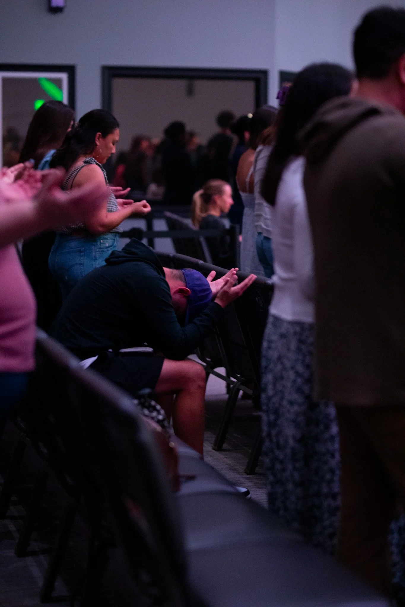 People attending a religious or spiritual service with many individuals standing and some with their eyes closed or hands raised in prayer.