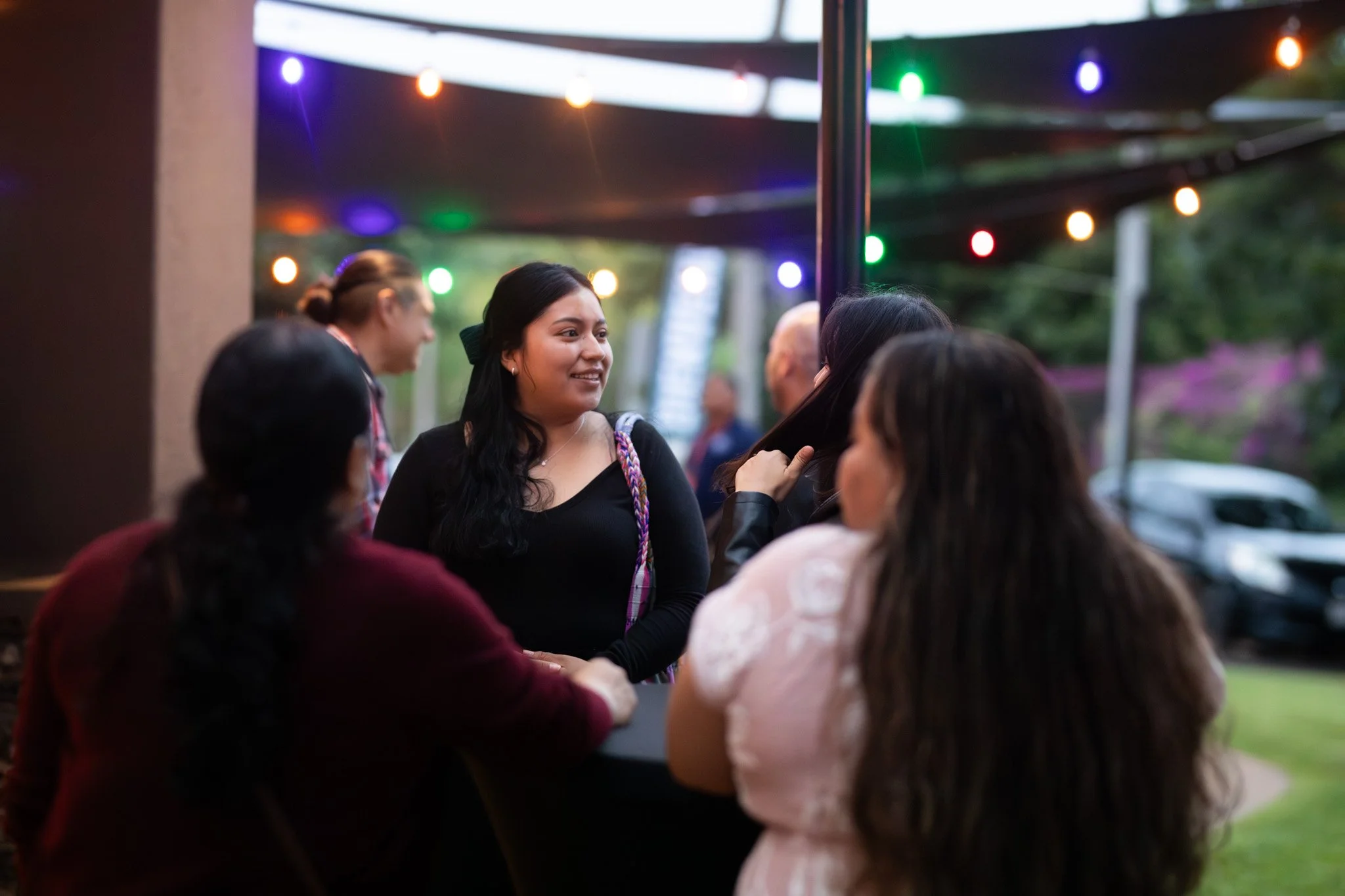 A group of women at an outdoor evening social event with colorful string lights, engaged in conversation.