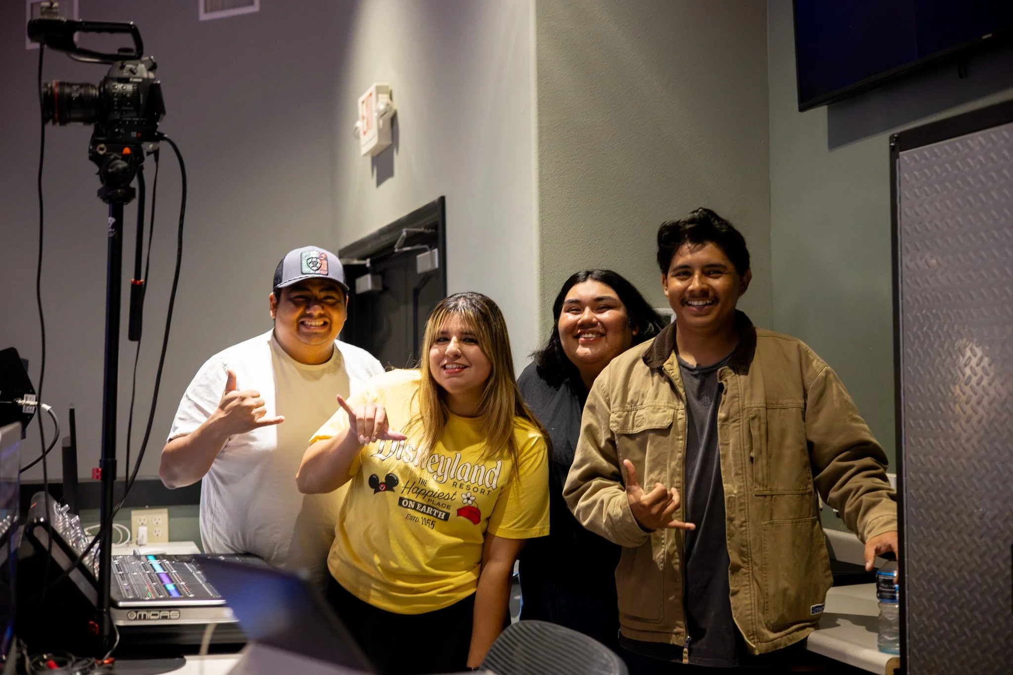Four young people in a room, smiling and making hand gestures, with audio equipment and a camera in the background.