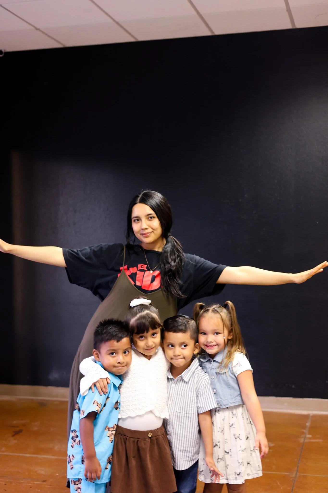 A person with short dark hair and a black t-shirt sitting at a table with a young girl with curly brown hair in a white polka dot shirt, in a room with a white wall and some black chairs.