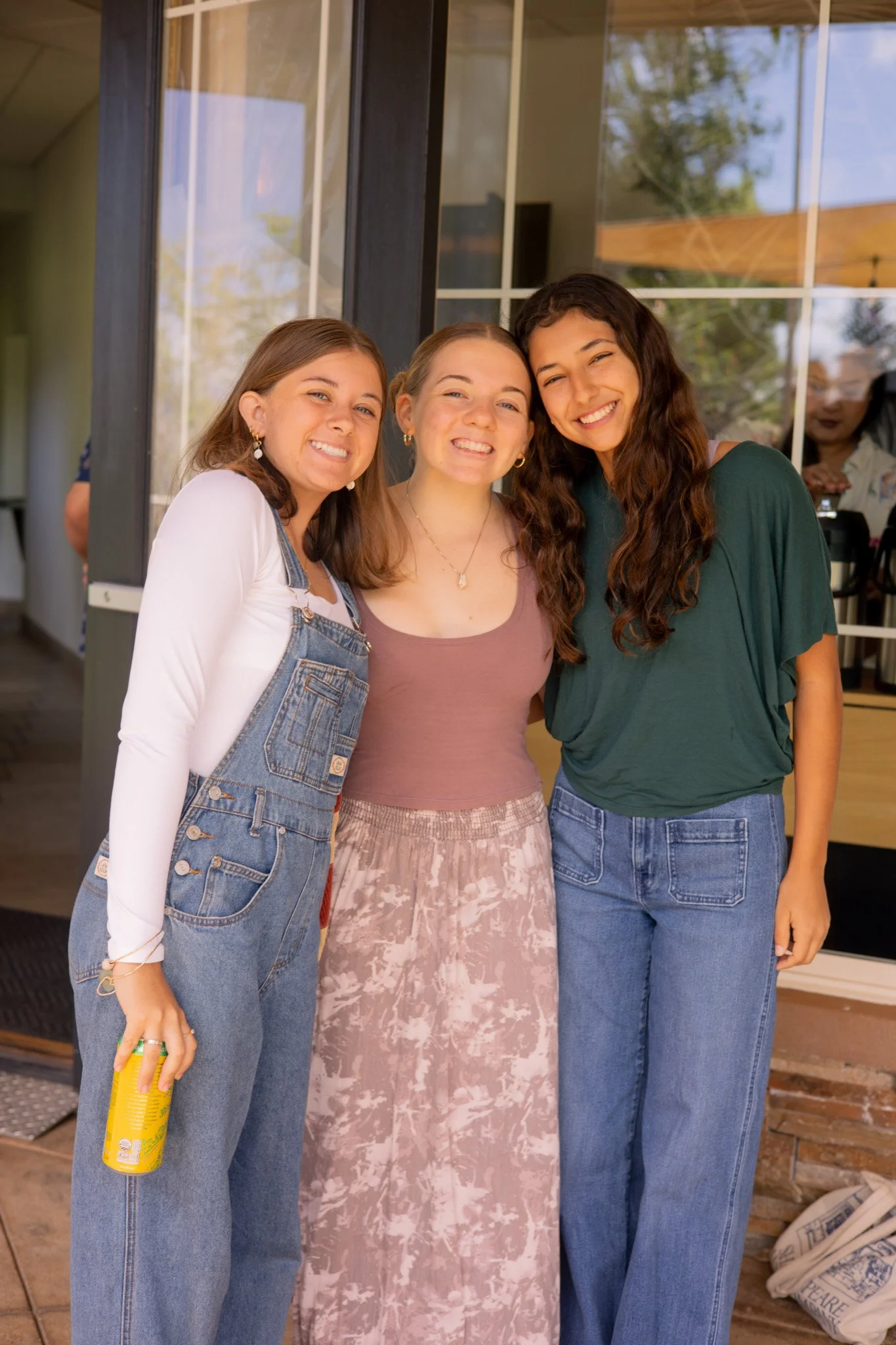 Three young women smiling and standing close together outside a building, with a large window behind them.