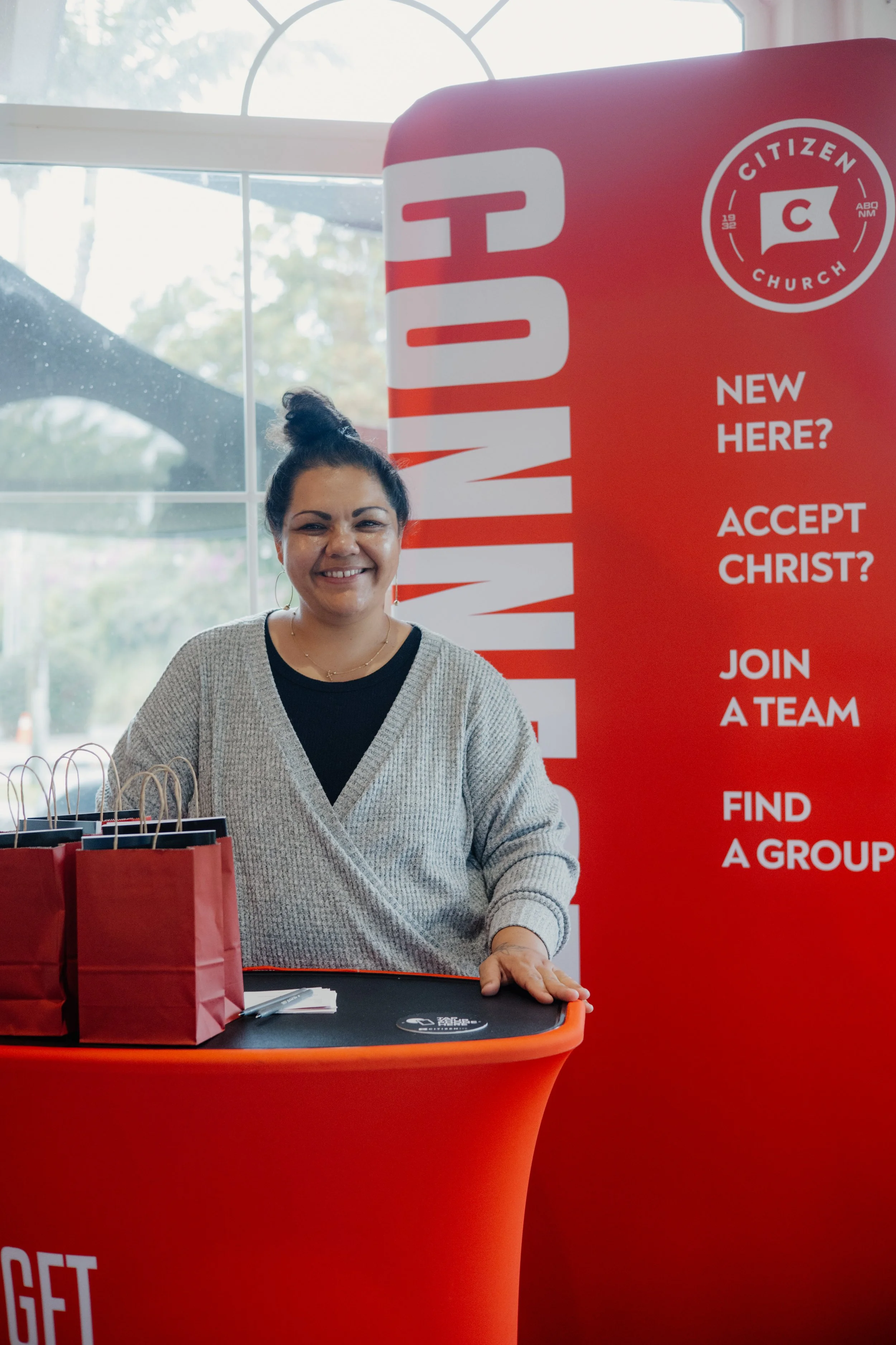 A woman with dark hair tied up in a bun smiling at a booth with Citizen Church branding, positioned next to a red banner that reads 'New here? Accept Christ? Join a team. Find a group.' on a sunny day inside a building with large windows.