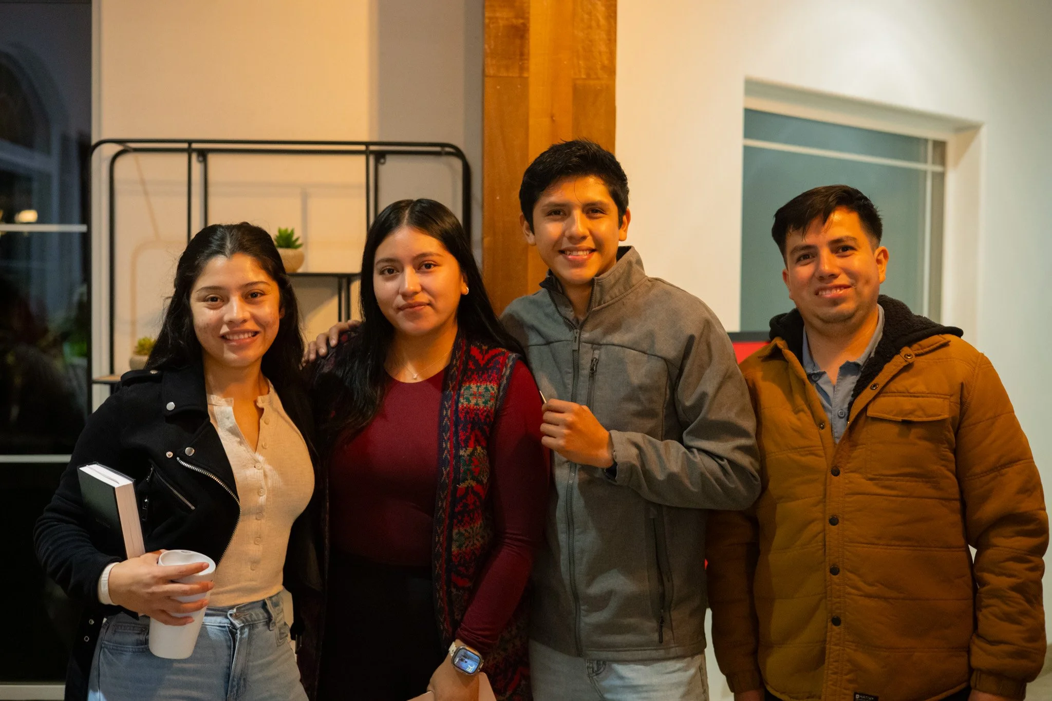 Four young adults standing close together indoors smiling at the camera, with a shelf and window in the background.