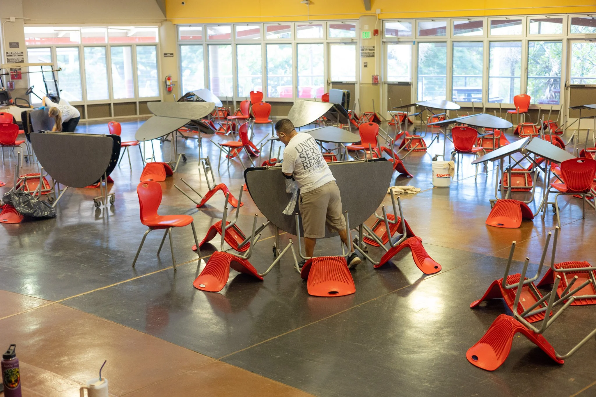 People cleaning and moving red chairs and gray tables in a large room with large windows and yellow walls.